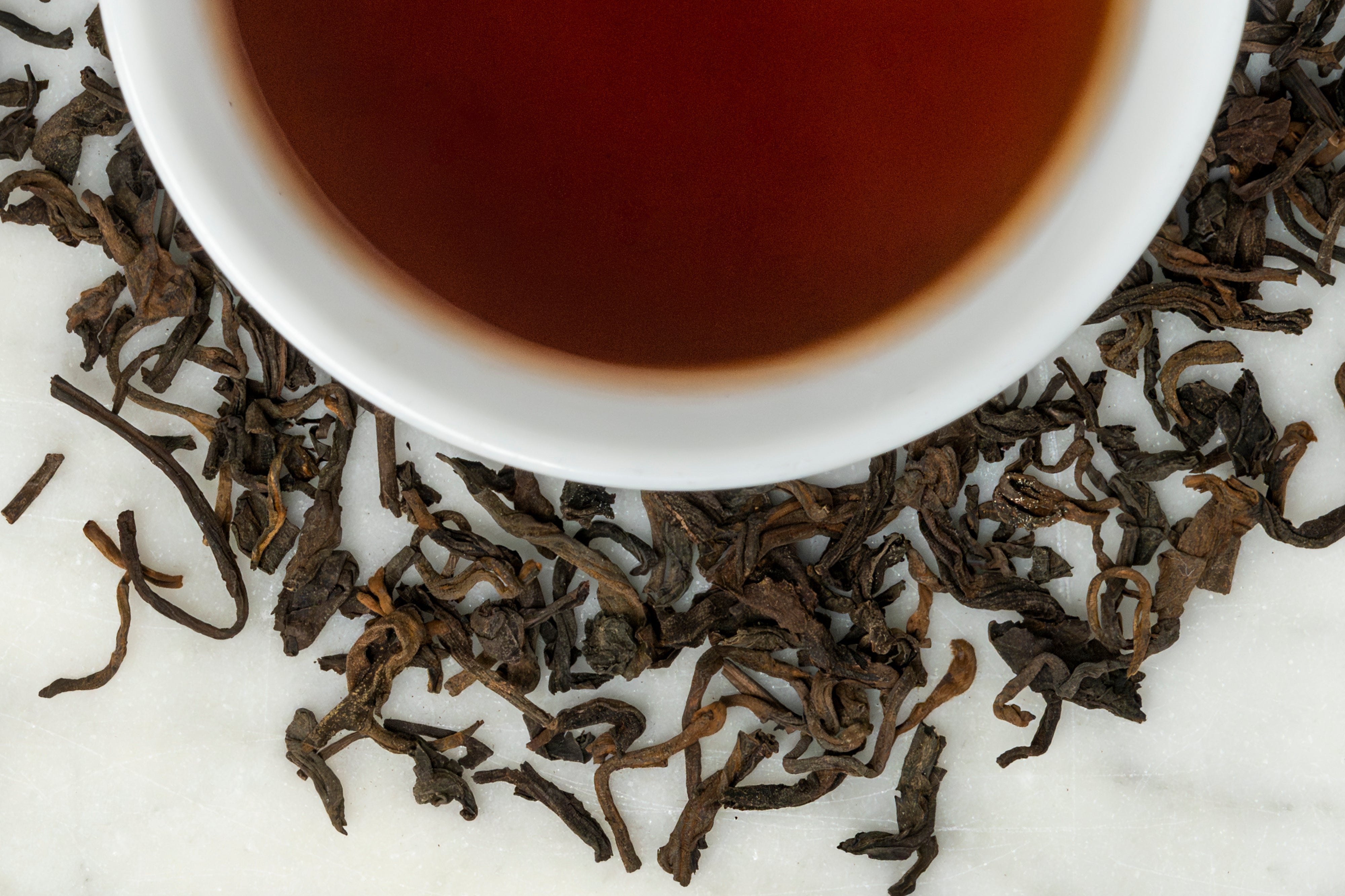 2016 Jingmai leaves and brewed tea. Dark brown twisted leaves surround a cup of dark, reddish-brown brewed tea.