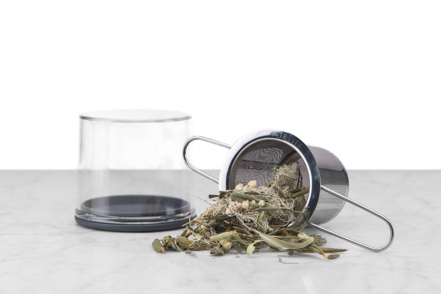 stainless steel tea infuser with folding handles staged with clear carrying case and spilling loose leaf tea on a marble and white background
