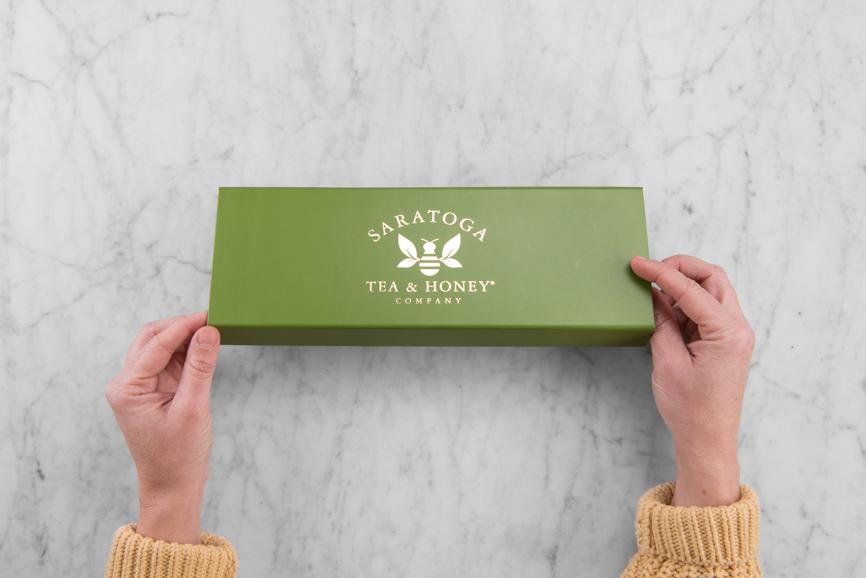 hands opening an oblong green gift box with a gold foil saratoga tea and honey co label