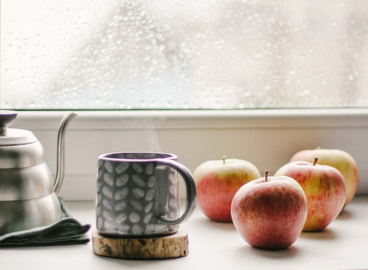 A steaming mug of tea sits next to a kettle and a few apples.