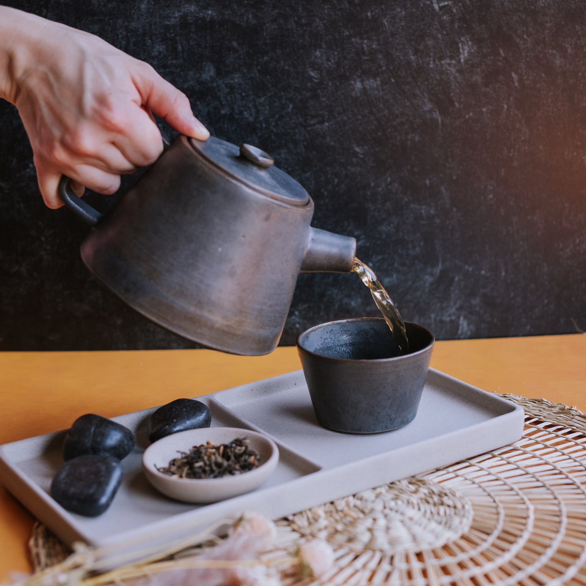 hand pouring black tea from ceramic teapot into a handleless modern teacup beside loose leaf black tea