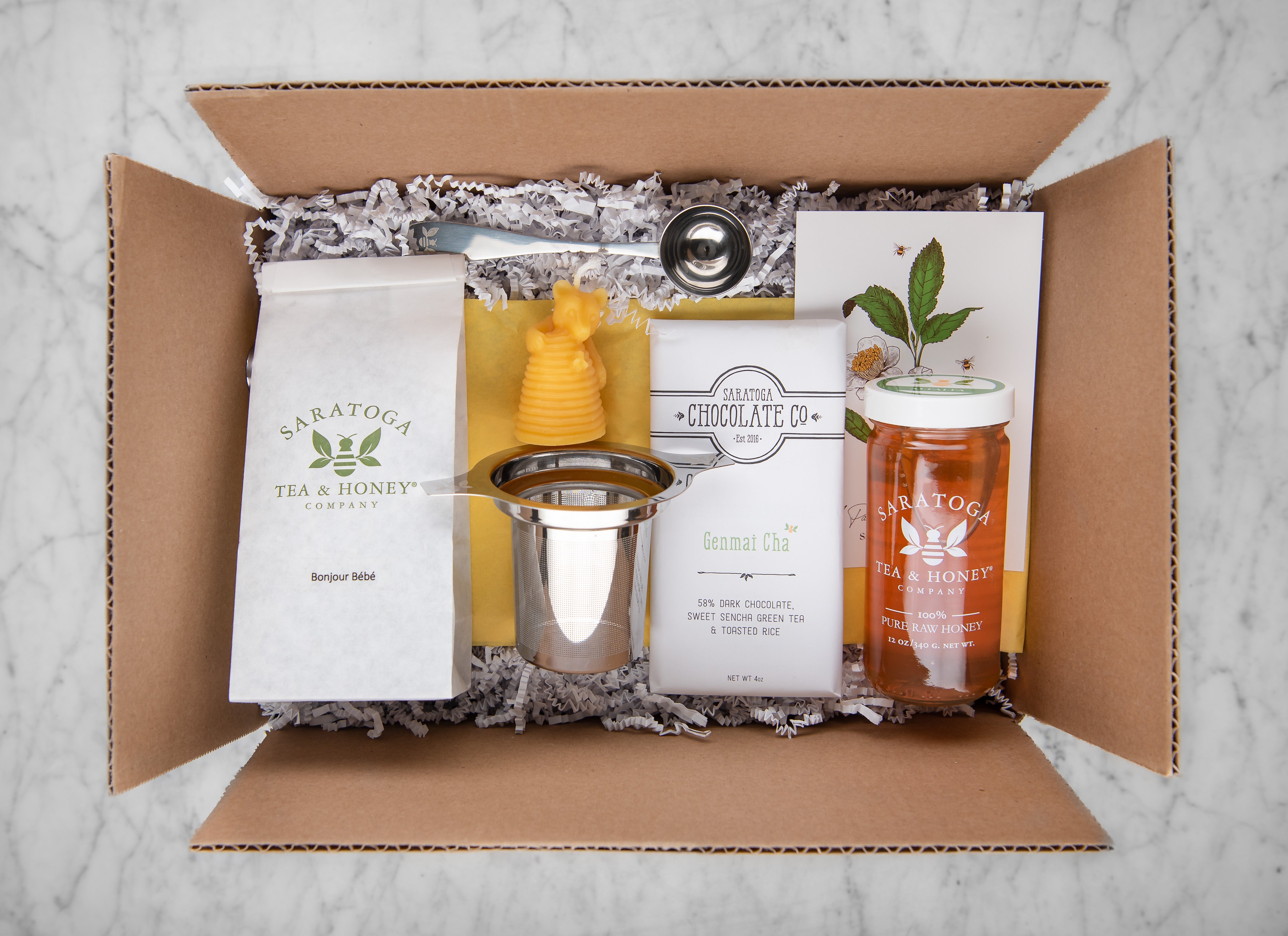 new baby tea and honey gift set featuring bonjour bebe breast feeding tea, alfalfa honey, a tea infuser, tea scoop, bear beeswax candle, and dark chocolate bar