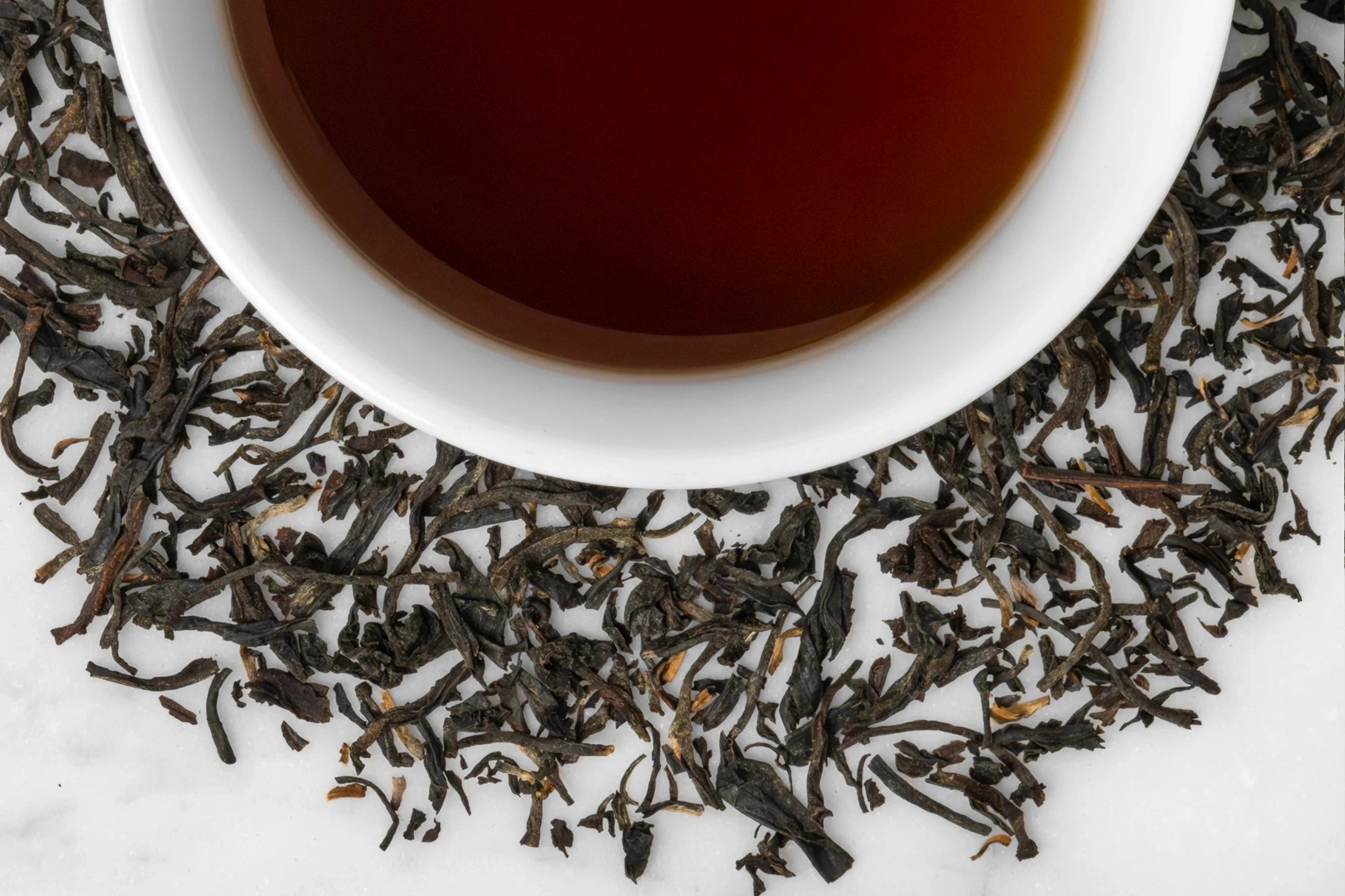 Robust Whole Assamica Black Tea Leaves Surround a Cup Of Dark Red Tea Liquor