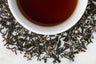 Robust Whole Assamica Black Tea Leaves Surround a Cup Of Dark Red Tea Liquor