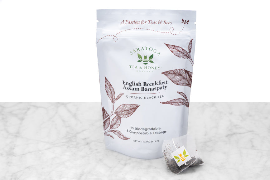 English Breakfast tea sachets packaging and example sachet. A small pyramid tea sachet with visible black tea leaves sits next to a pouch reading "English Breakfast Assam Banaspaty Organic Black Tea. 15 Biodegradable & Compostable Tea Bags."