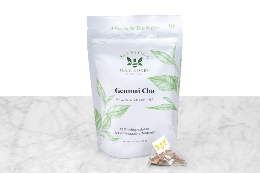 Genmai Cha green tea sachets packaging and example sachet. A small pyramid tea sachet with visible green tea leaves and brown toasted rice sits next to a pouch reading "Genmai Chai Organic Green Tea. 15 Biodegradable & Compostable Tea Bags."