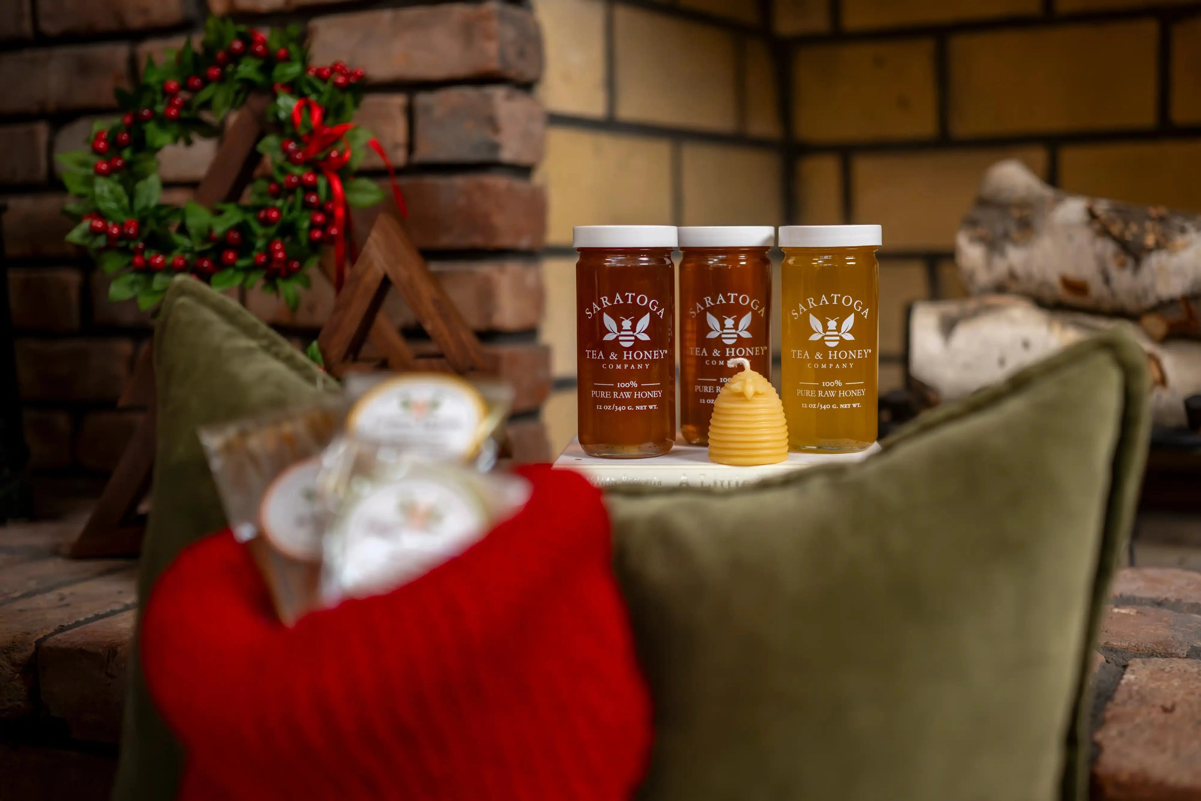 In focus: 3 Saratoga Tea & Honey Co. jars of raw honey next to a beeswax beehive candle. Blurred in foreground: stocking with mini honey candy stocking stuffers