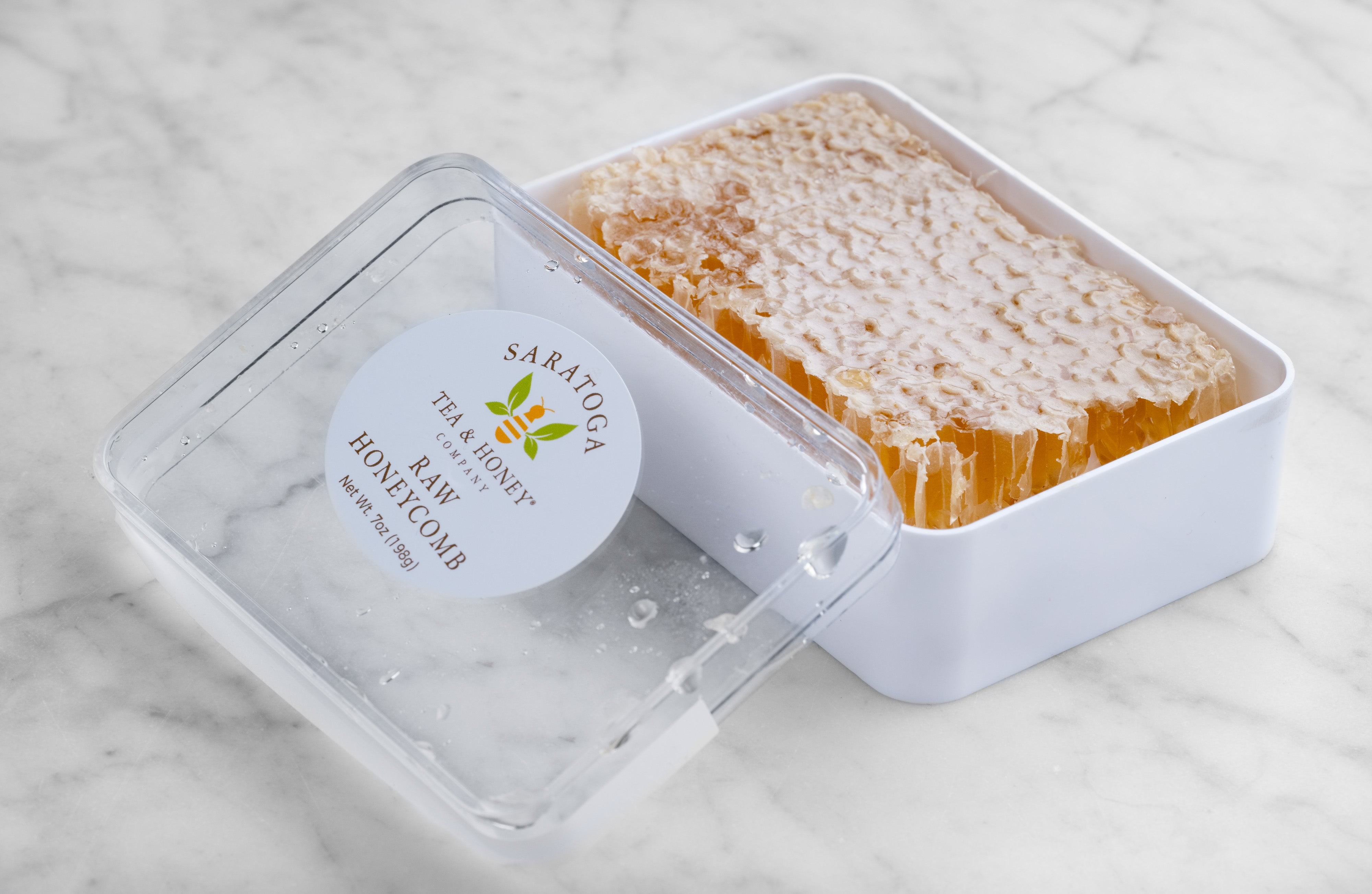 A rectangle of golden acacia honeycomb sits in an open white box. A clear lid with a sicker reading "Raw Honeycomb" is propped to the left side of the honeycomb and box.