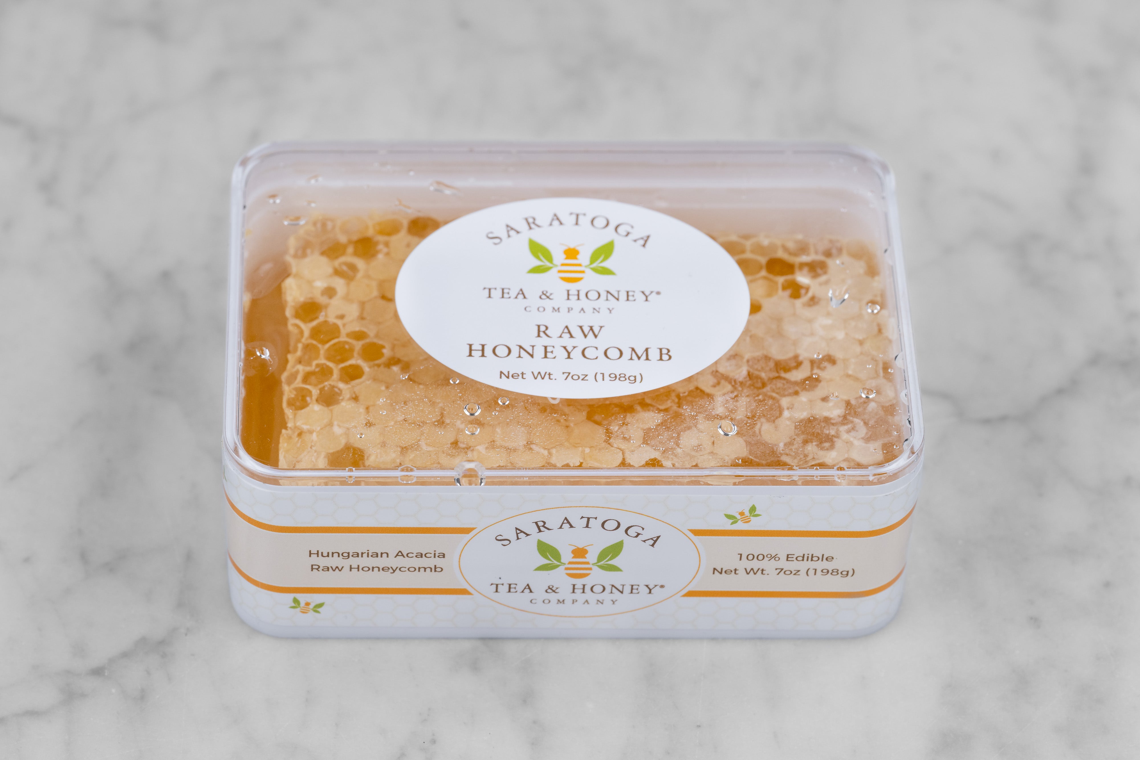 A rectangle of golden Acacia honeycomb in a white box with a clear lid, seen from a side-top angle. The top sticker displays the logo and reads: "Raw Honeycomb. Net Wt. 7oz). The side sticker reads "Hungarian Acacia Raw Honeycomb. 100% Edible."