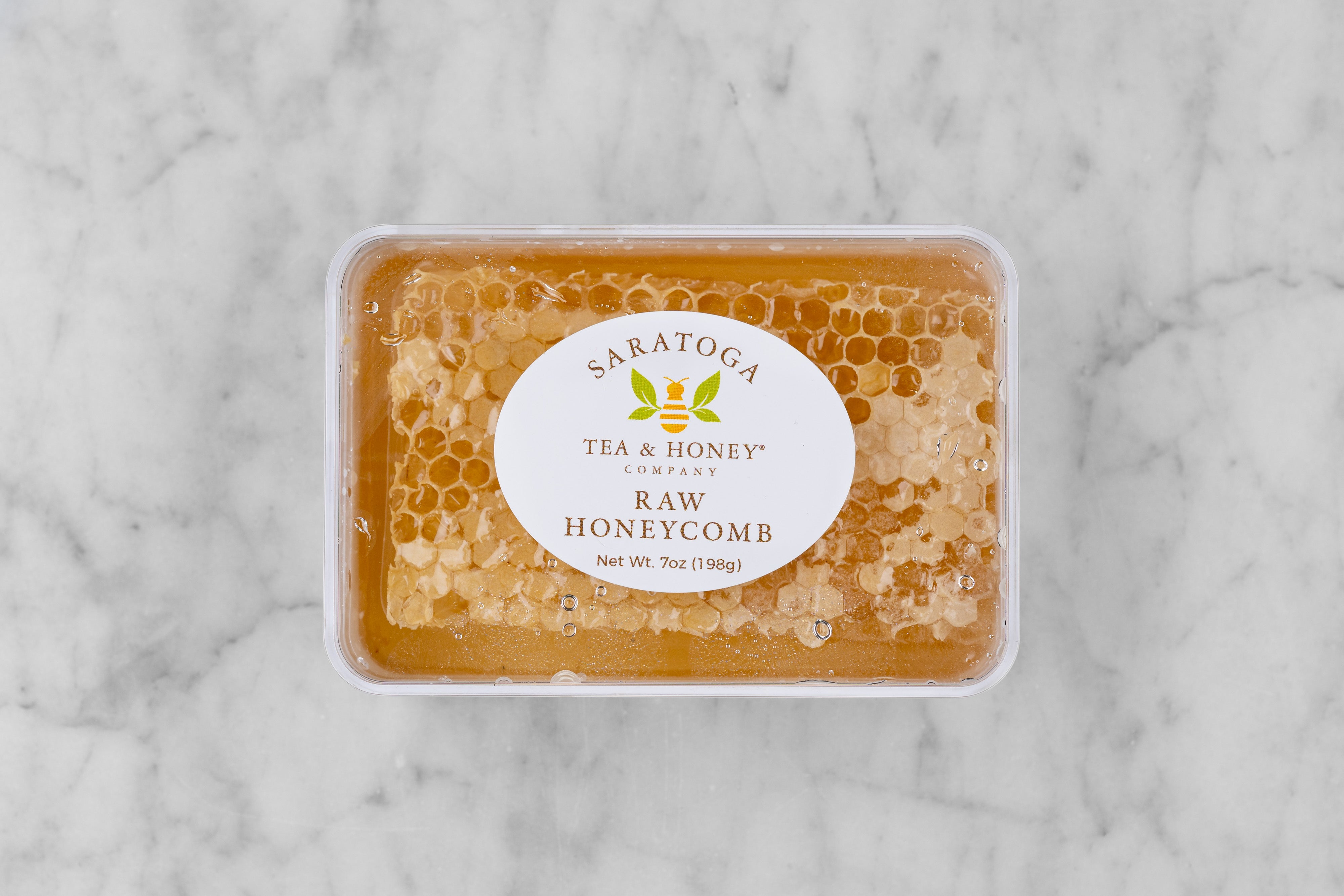 A rectangle of golden honeycomb in a white box with a clear lid seen from above. The lid has a sticker with the logo and reads "Raw Honeycomb". 