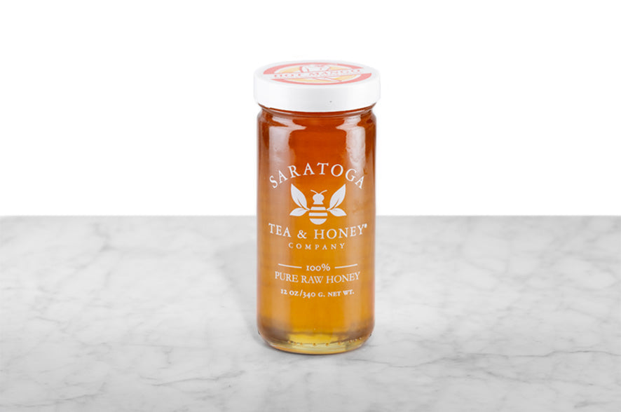 Hot Mango Honey displayed in a 12oz cylindrical glass jar with a white lid. The honey fades in color from a rich, reddish amber at the top to a sunny gold at the bottom of the jar. The logo and the words: "100% Pure Raw Honey" are printed on the jar. 