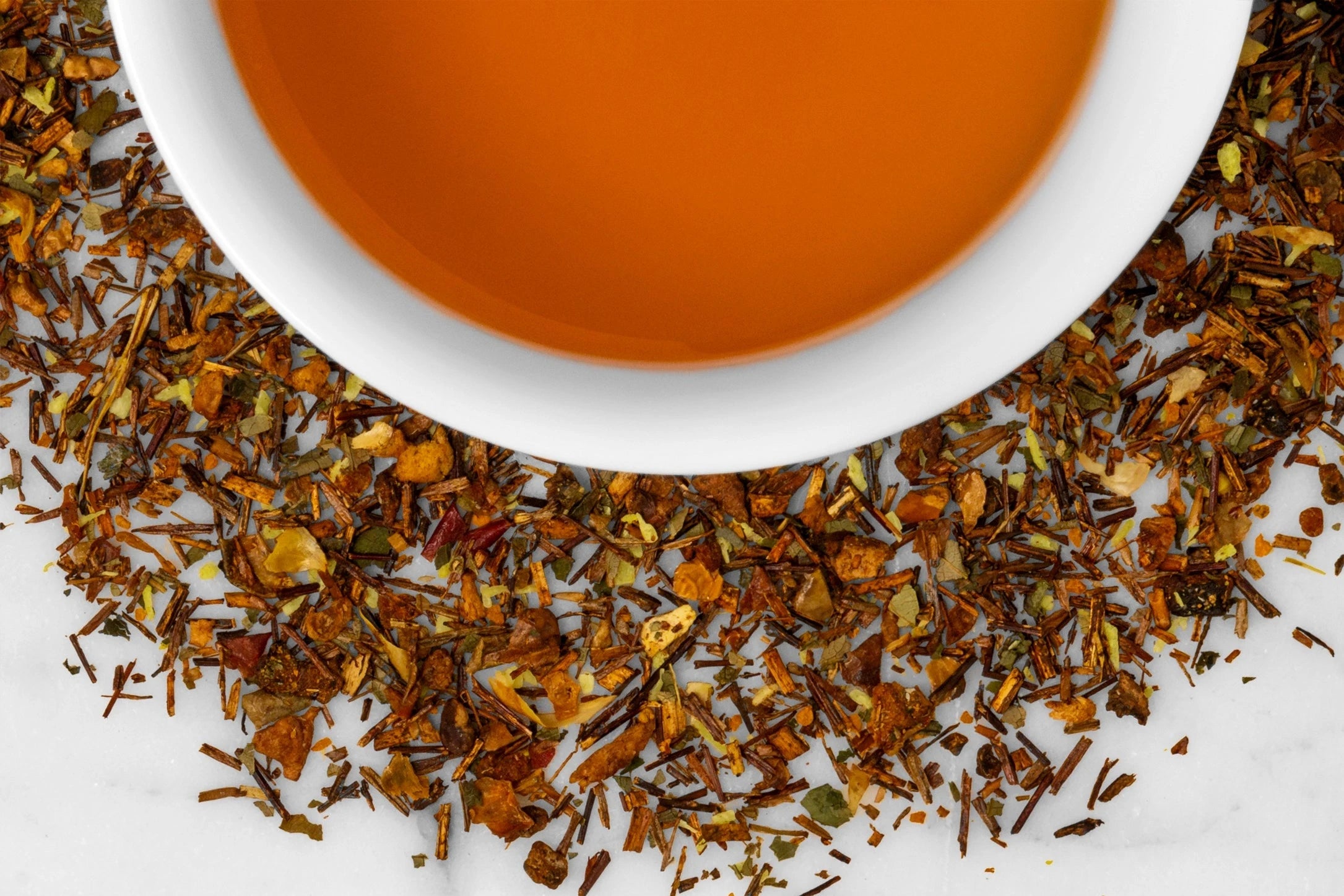 Close up of dry and brewed Island Glow. The dry tea is composed of rooibos with turmeric, mango, and coconut pieces surrounding a cup of brewed, orange-hued tea.