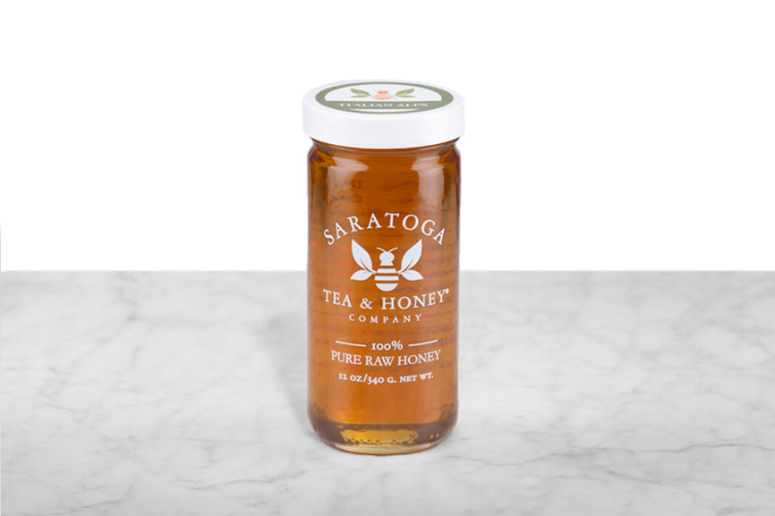 Amber-hued Italian Alps Honey displayed in a 12oz cylindrical glass jar with a white lid. The logo and the words: "100% Pure Raw Honey" are printed on the jar.