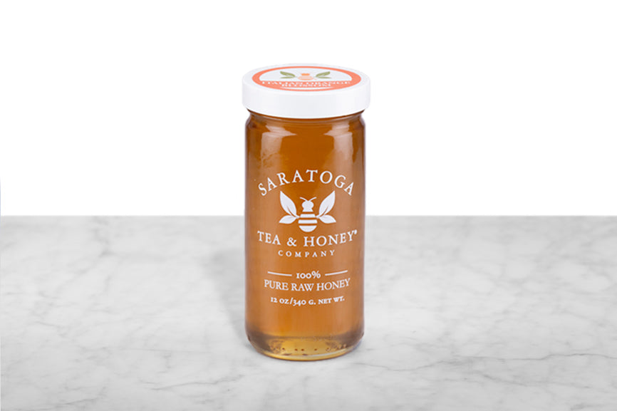 Light amber-hued Italian Orange Blossom Honey displayed in a 12oz cylindrical glass jar with a white lid. The logo and the words: "100% Pure Raw Honey" are printed on the jar.