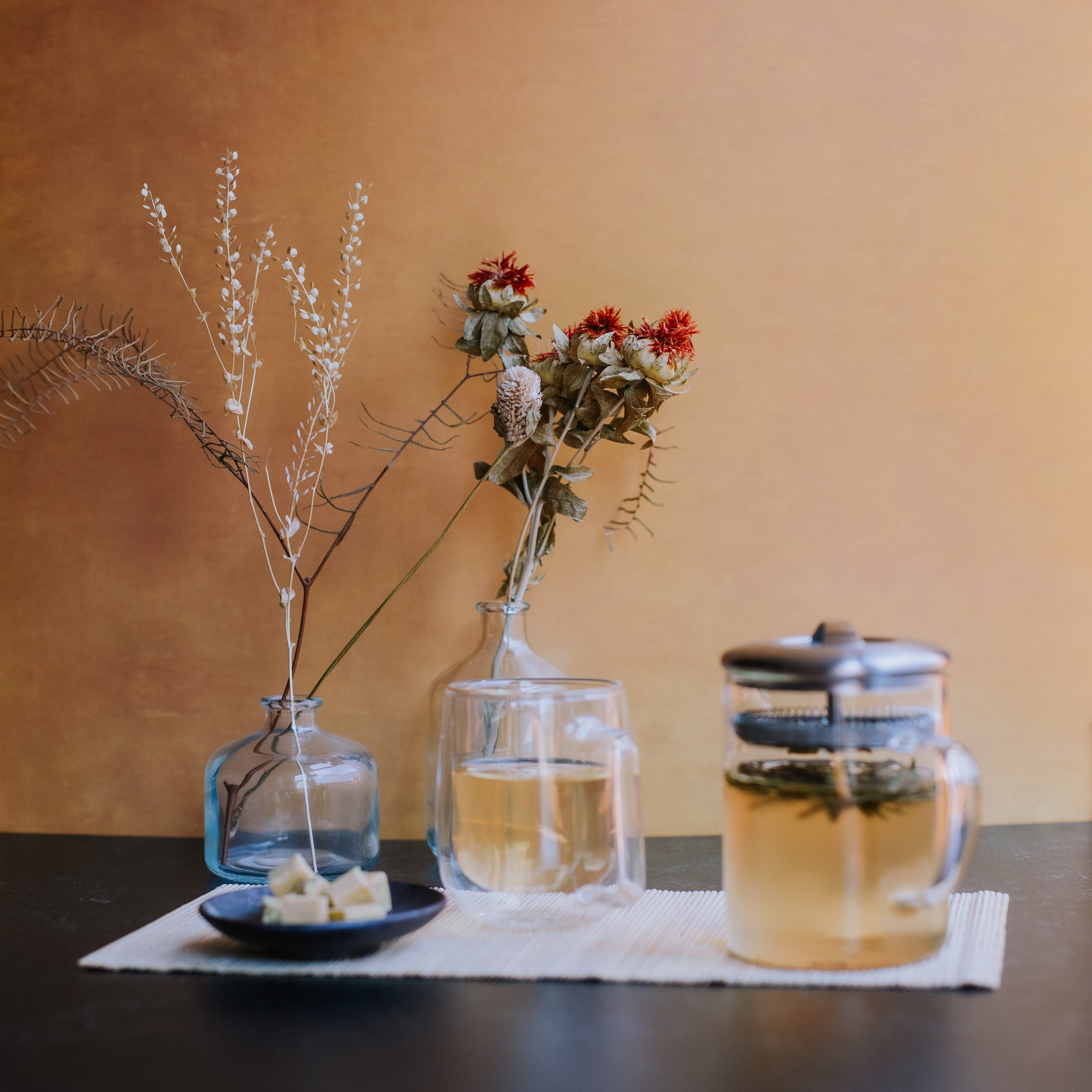 green tea brewing in a glass container with a glass mug of green tea and dish of mung bean cake against a mustard yellow background with dried flowers