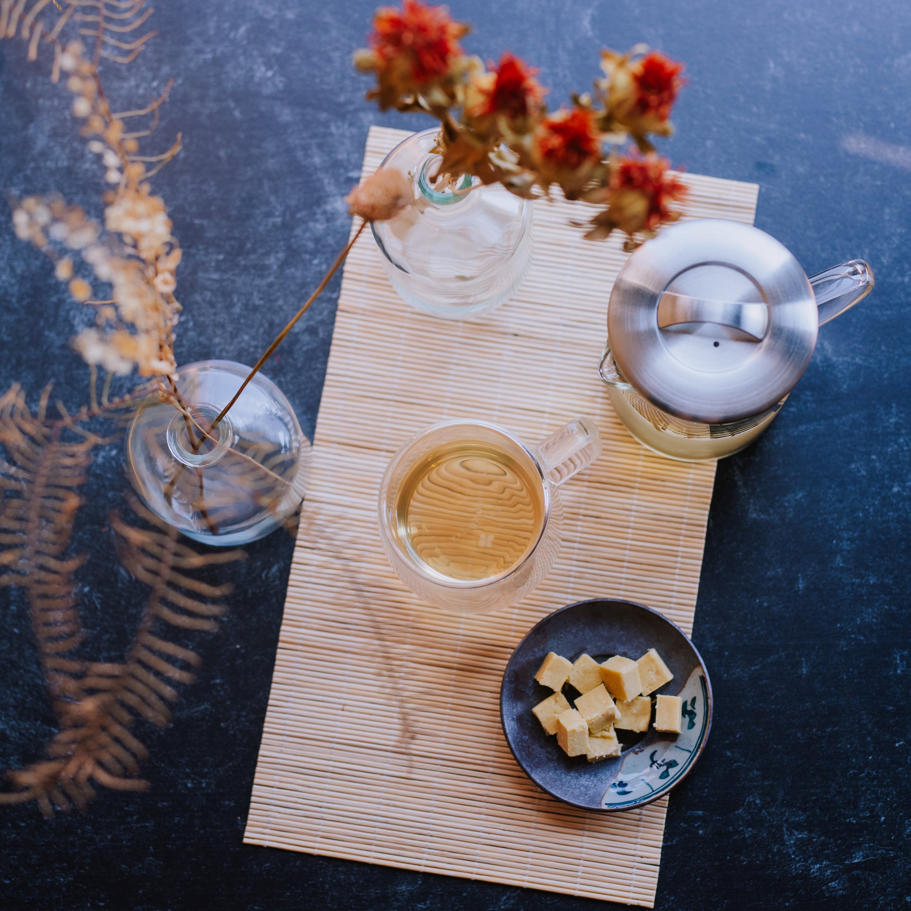 Aesthetic display of brewed green tea alongside a glass teapot, plate of mung bean cake squares, and dried florals.