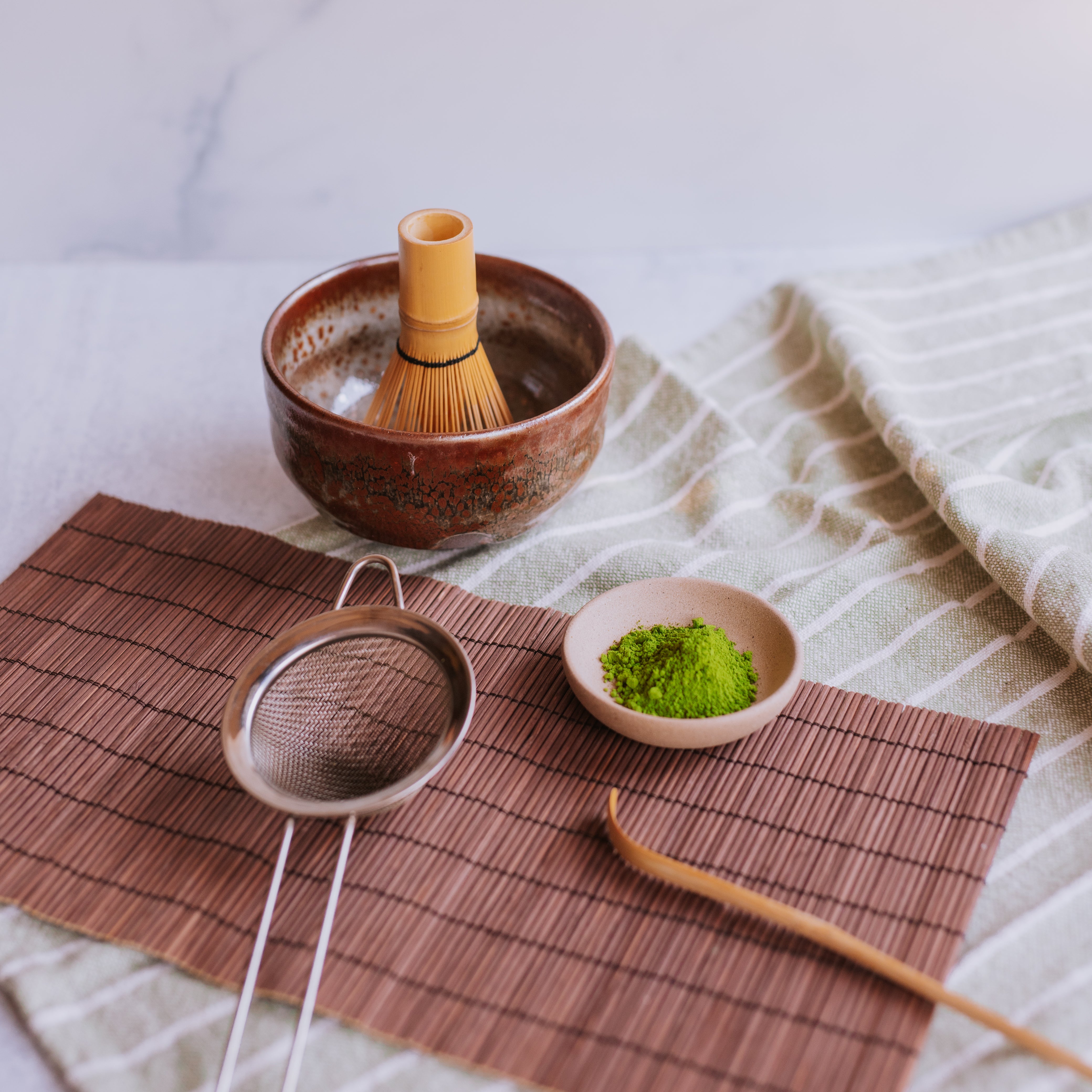 flat lay photo of brown glaze matcha bowl (chawan), match whisk (chasen), bamboo scoop (chashuka), stainless steel sieve, and matcha powder