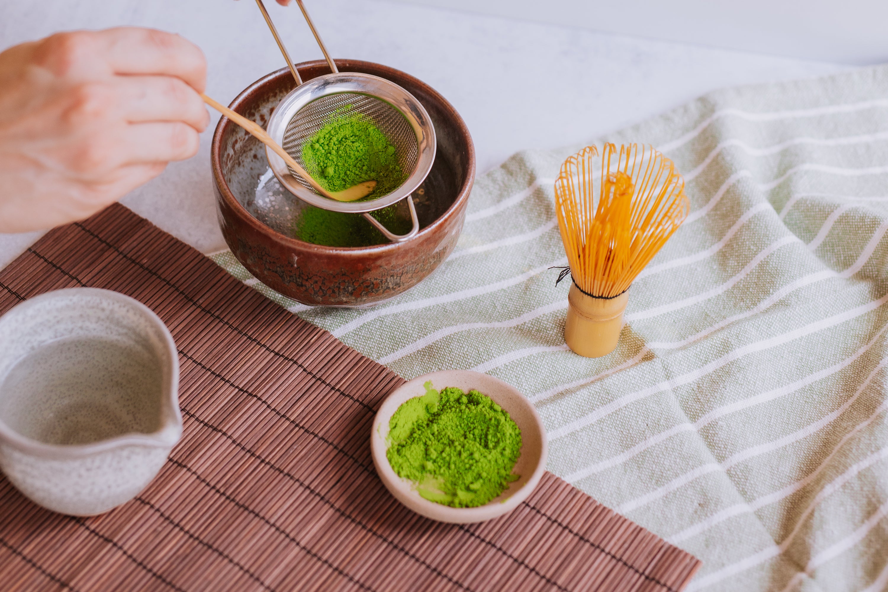 hand sifting matcha through a sieve into a matcha chawan with whisk, pitcher, and dish of matcha powder
