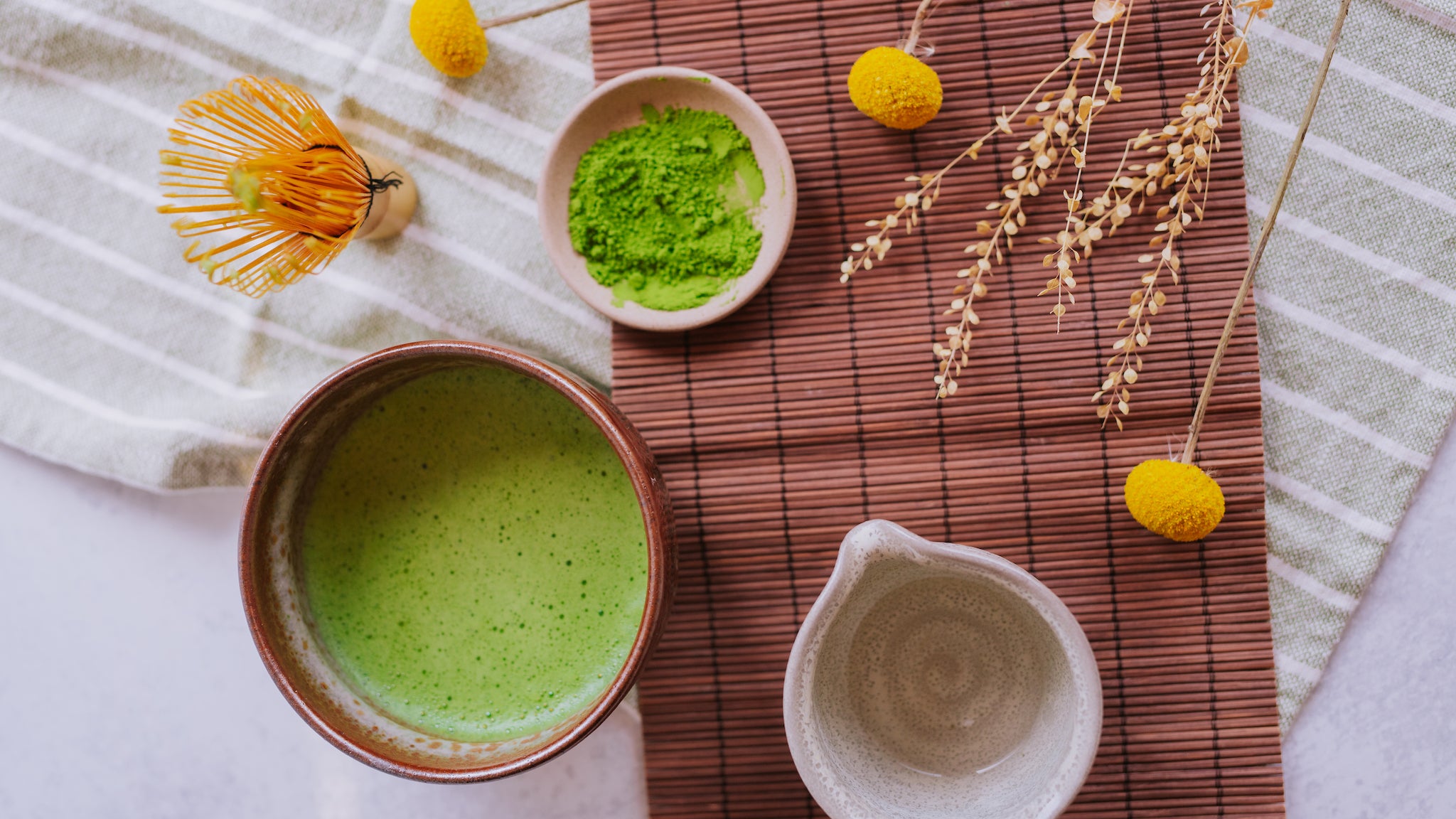 Bowl of matcha, matcha whisk (chasen), matcha powder, and small pitcher of water on a bamboo background