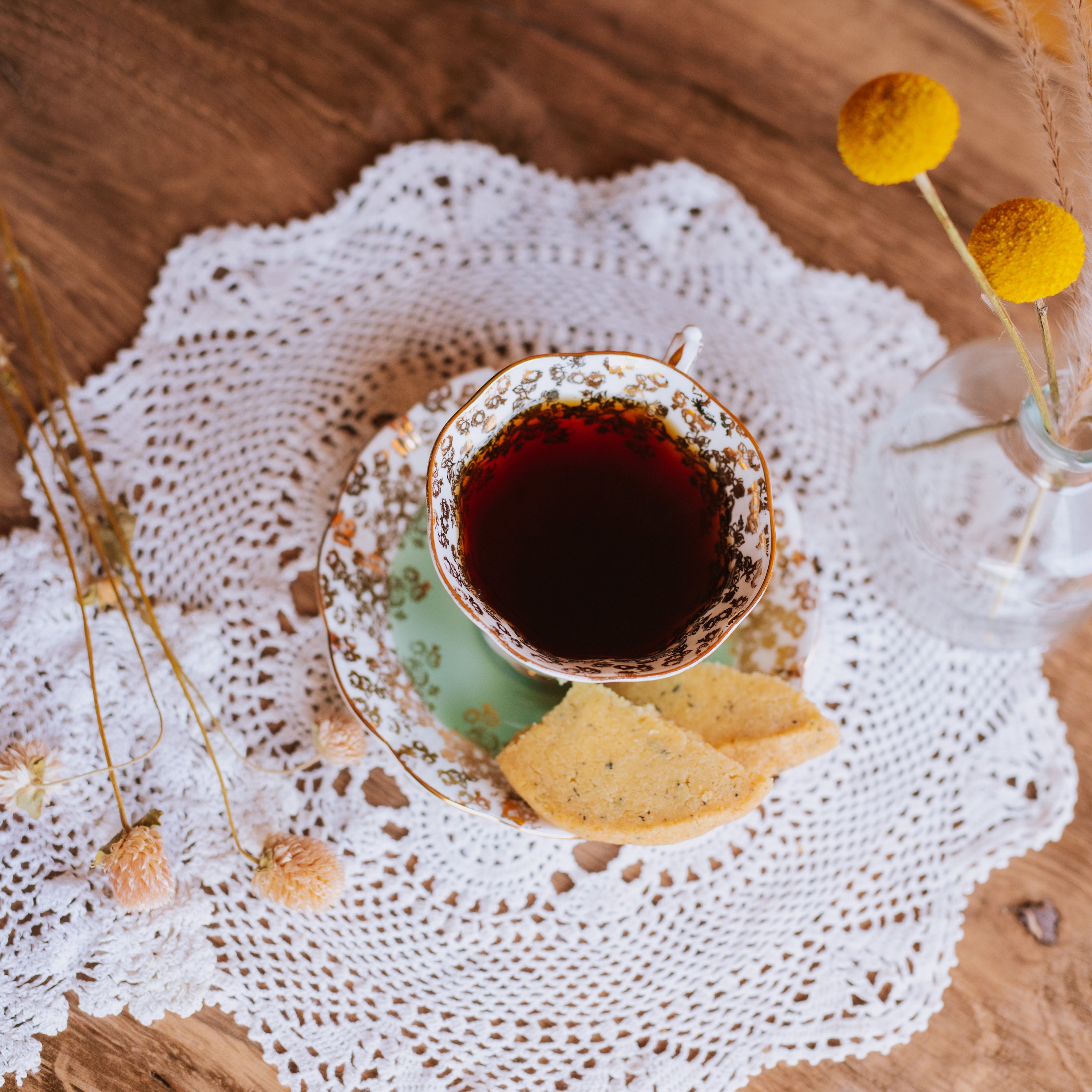 teal and gold flowered vintage teacup with black tea and shortbread cookies on the side arranged on a lace doily with dried flowers