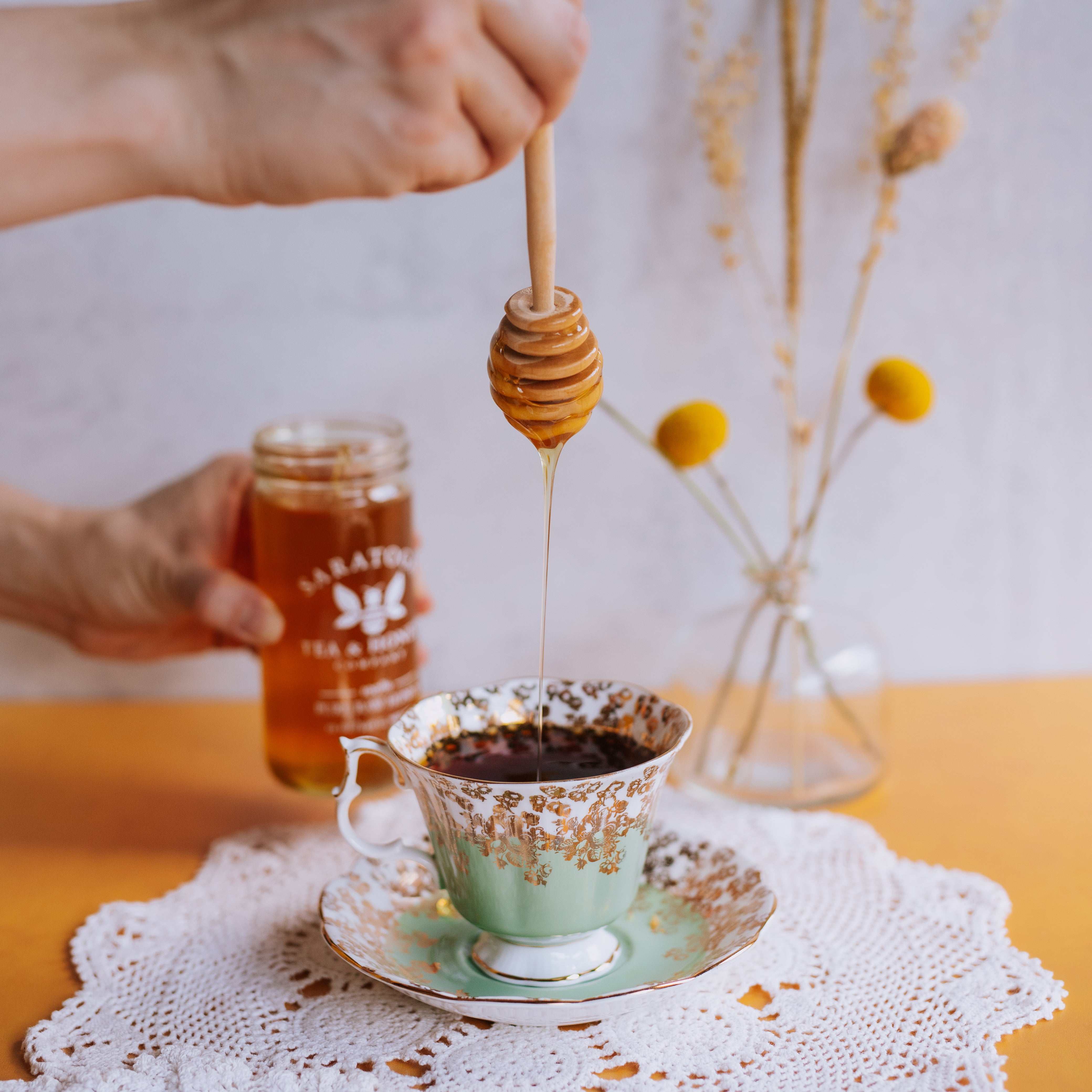 hand drizzling honey into vintage teacup from above with a wooden honey dipper and Saratoga Tea & Honey Co. honey jar in background