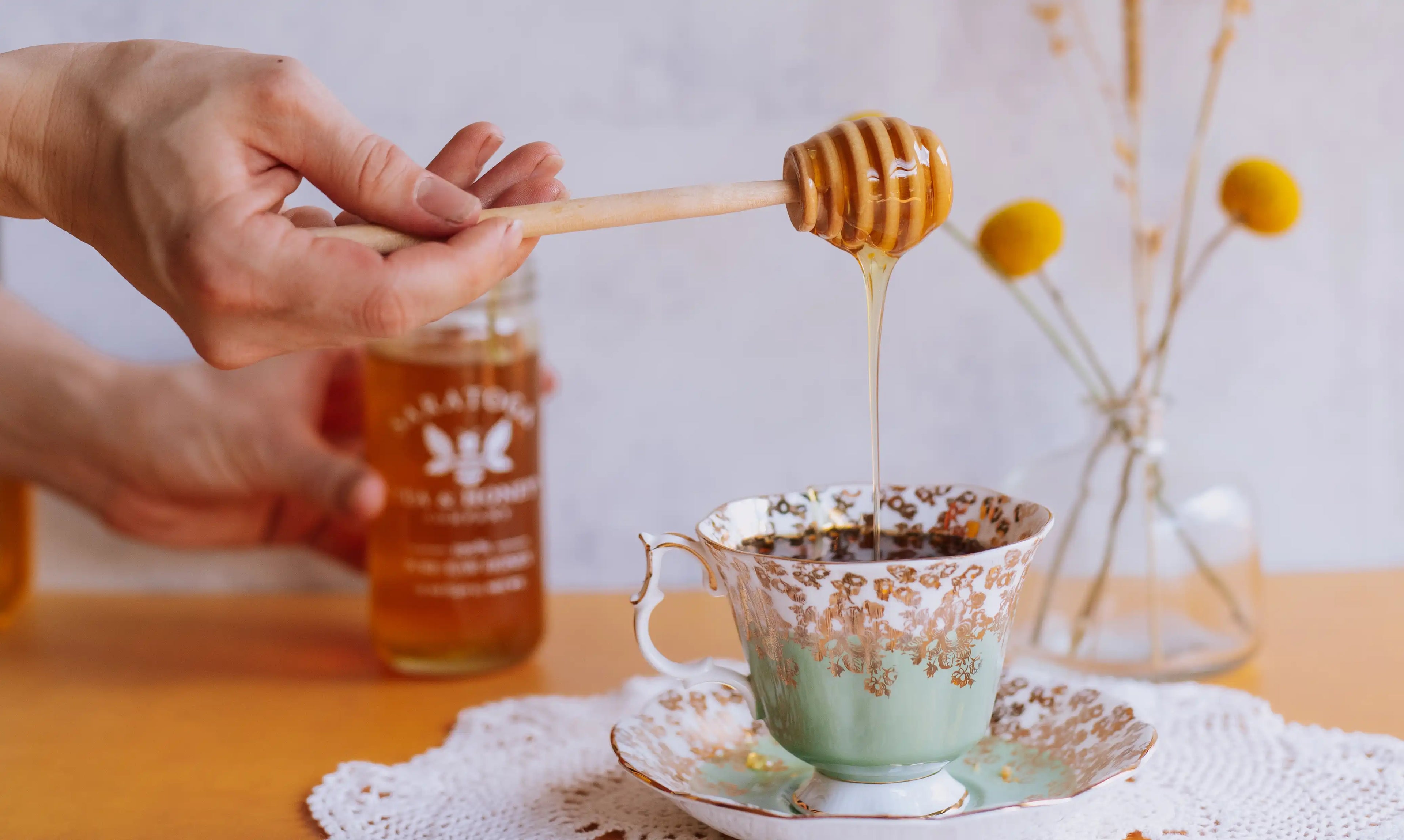 hand holding honey dipper drizzling honey into a mint colored vintage tea cup with jar of saratoga tea and honey co in the background