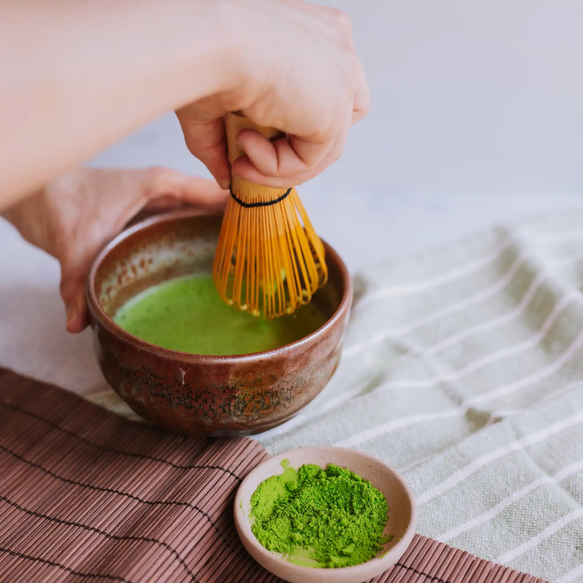 hand whisking a bowl of matcha with a bamboo whisk and a dish of matcha powder in the foreground