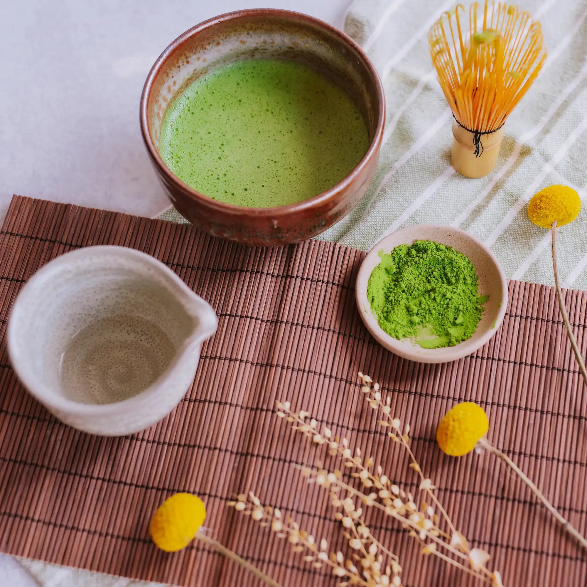 flay lay of whisked matcha in a hand-thrown matcha bowl with a dish of matcha powder on tatami mats with dried florals