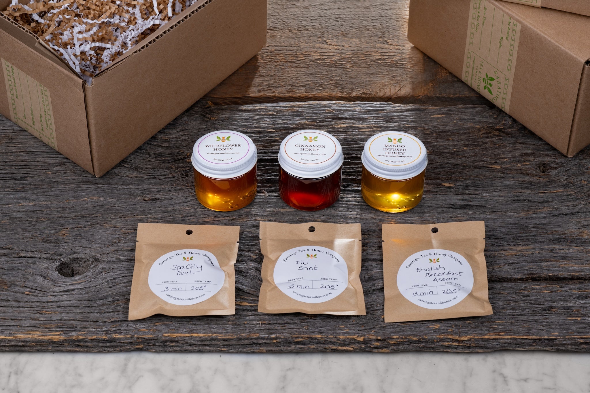 flat lay on wood background of three sample teas and three sample honeys with shipping boxes and decorative fill