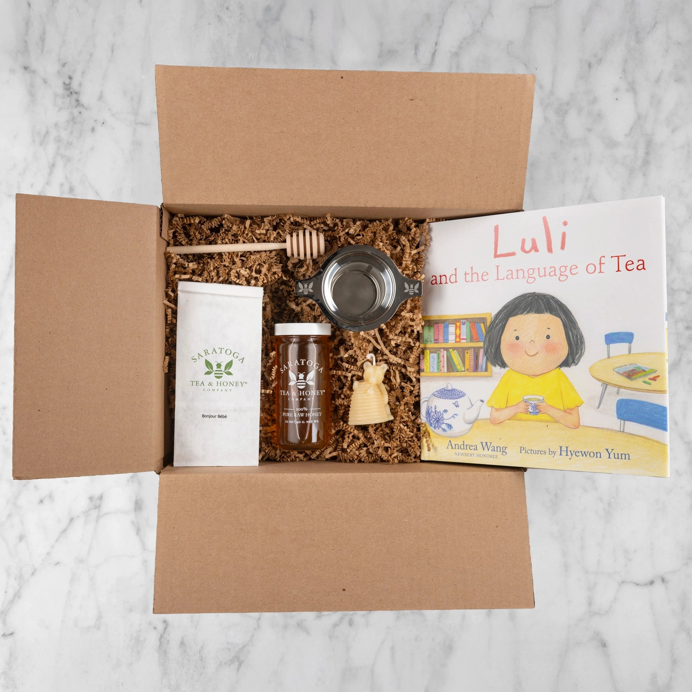 New Mom & Baby Bundle products in an open cardboard box with kraft crinkle fill: Bonjour Bébé tea in a white bag, Alfalfa Honey in a glass jar, wooden honey dipper, stainless steel tea infuser with bee etchings, small yellow beeswax bear and hive candle, and a picture book with the title Luli and the Language of Tea