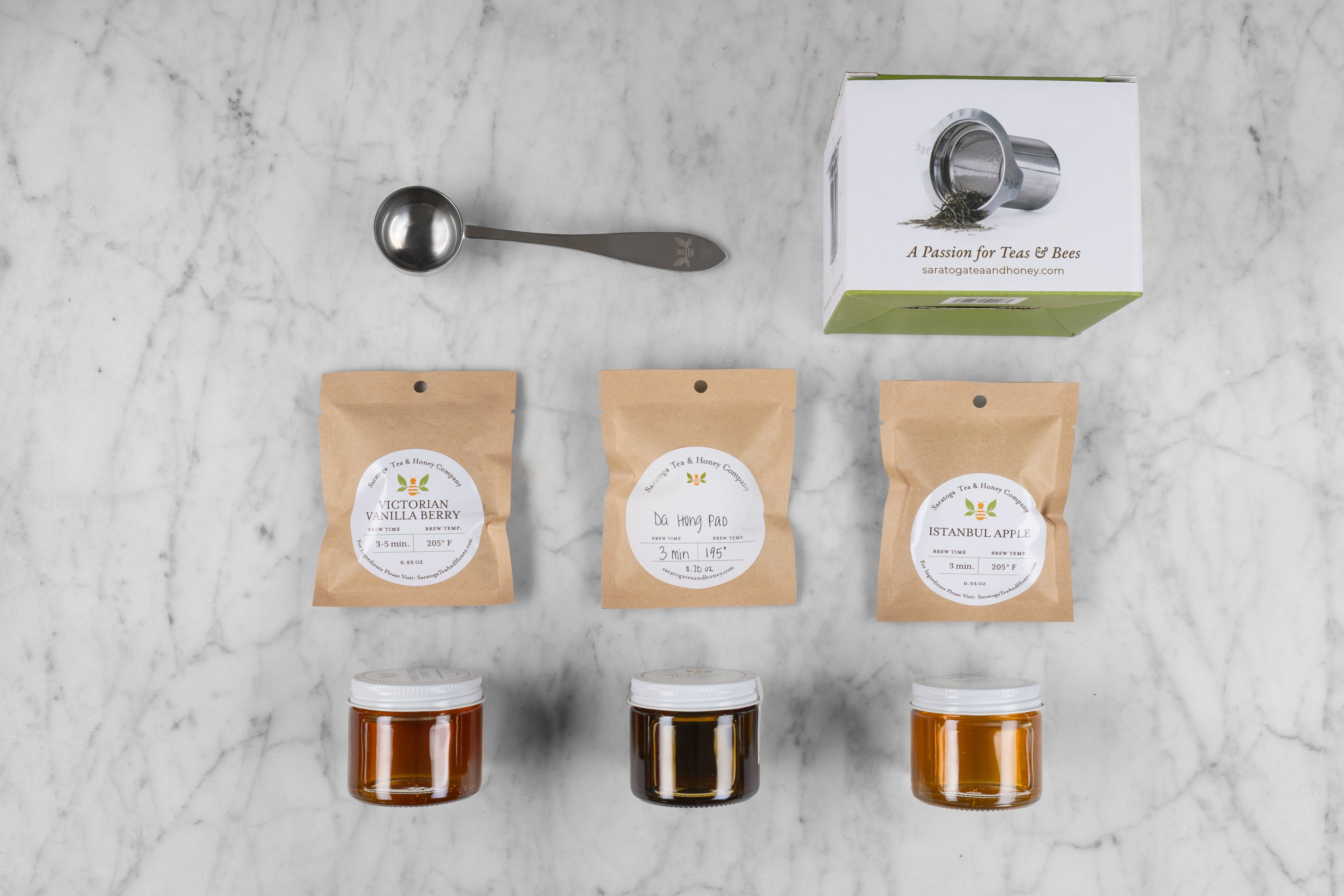 flat lay of new bee gift set with three mini honeys, 3 sample teas, tea scoop, and stainless steel tea infuser on a marble background