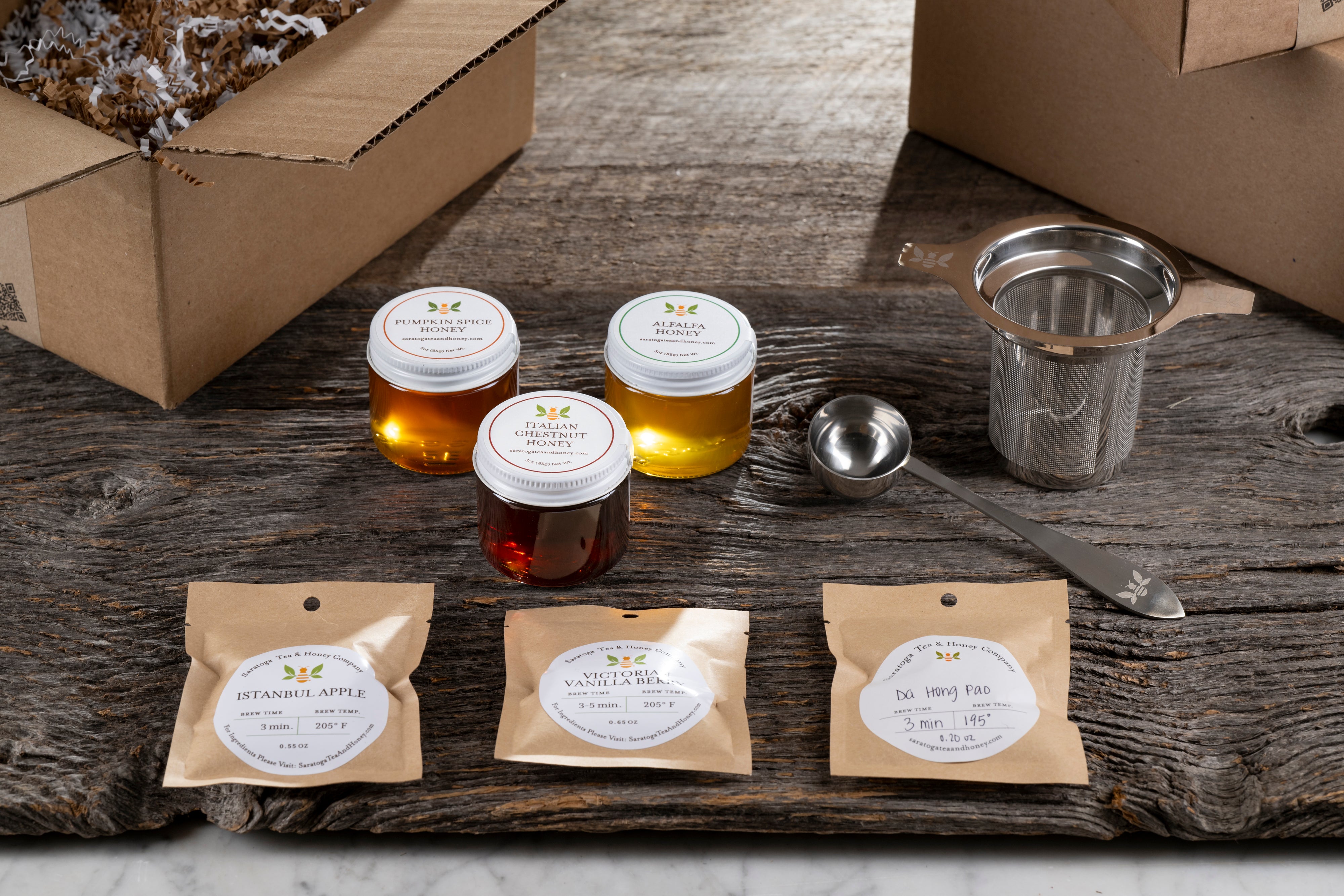 new bee gift set contents on wood with shipping boxes. includes: three mini honeys, three tea samples, stainless steel infuser, and tea scoop.