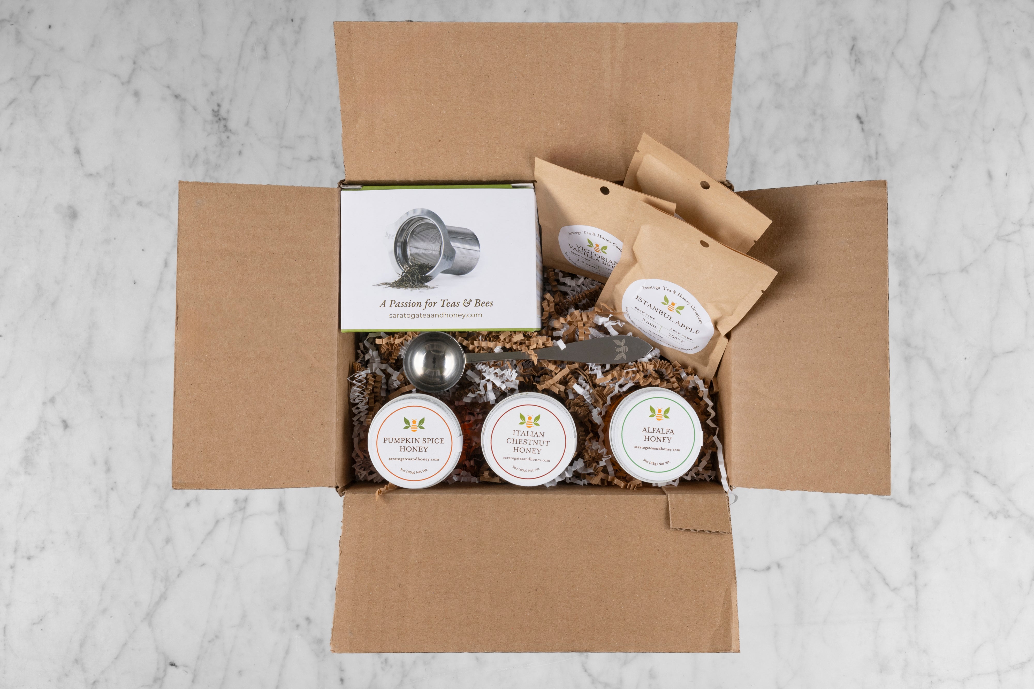 new bee gift set including three sample teas, 3 sample honeys, a stainless steel tea infuser, and a tea scoop in a shipping box with gift fill on marble background