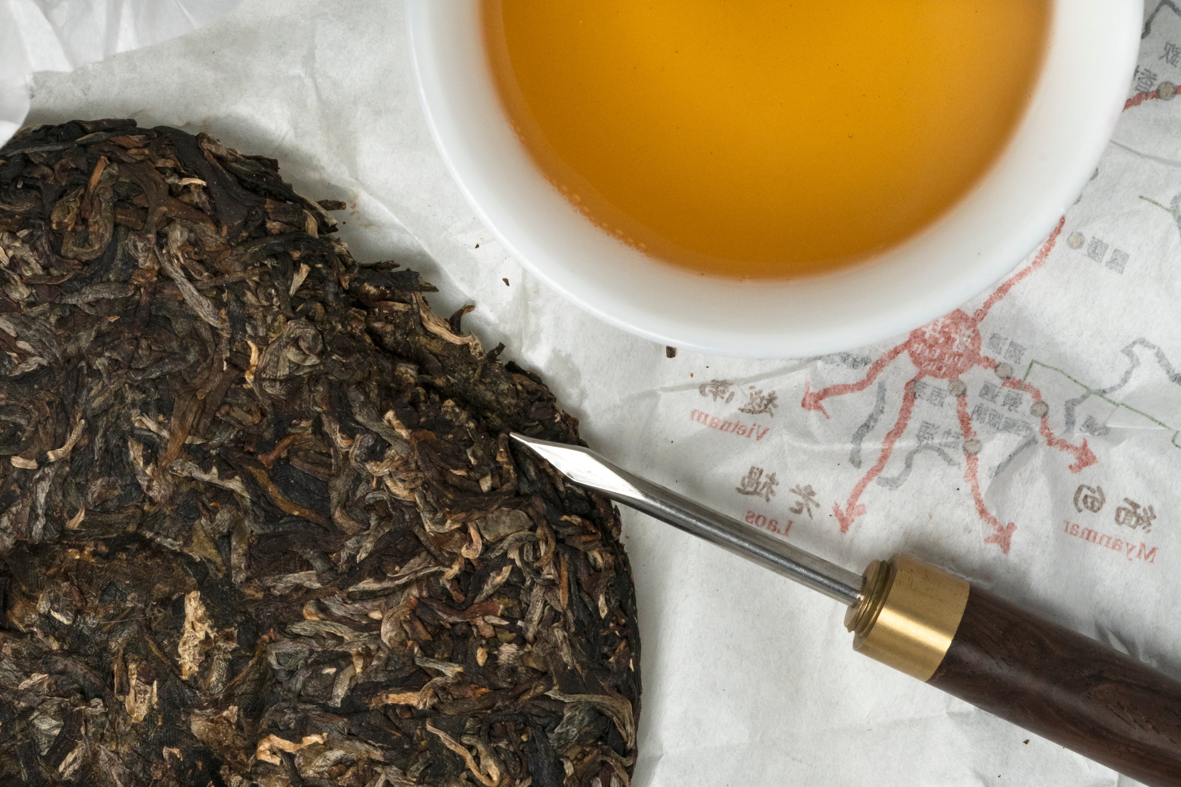 2014 Jingmai Old Forest tea cake and brewed tea. Unwrapped sheng pu erh pressed tea cake with a silver tea pick and a cup of old-gold-colored brewed pu erh tea.