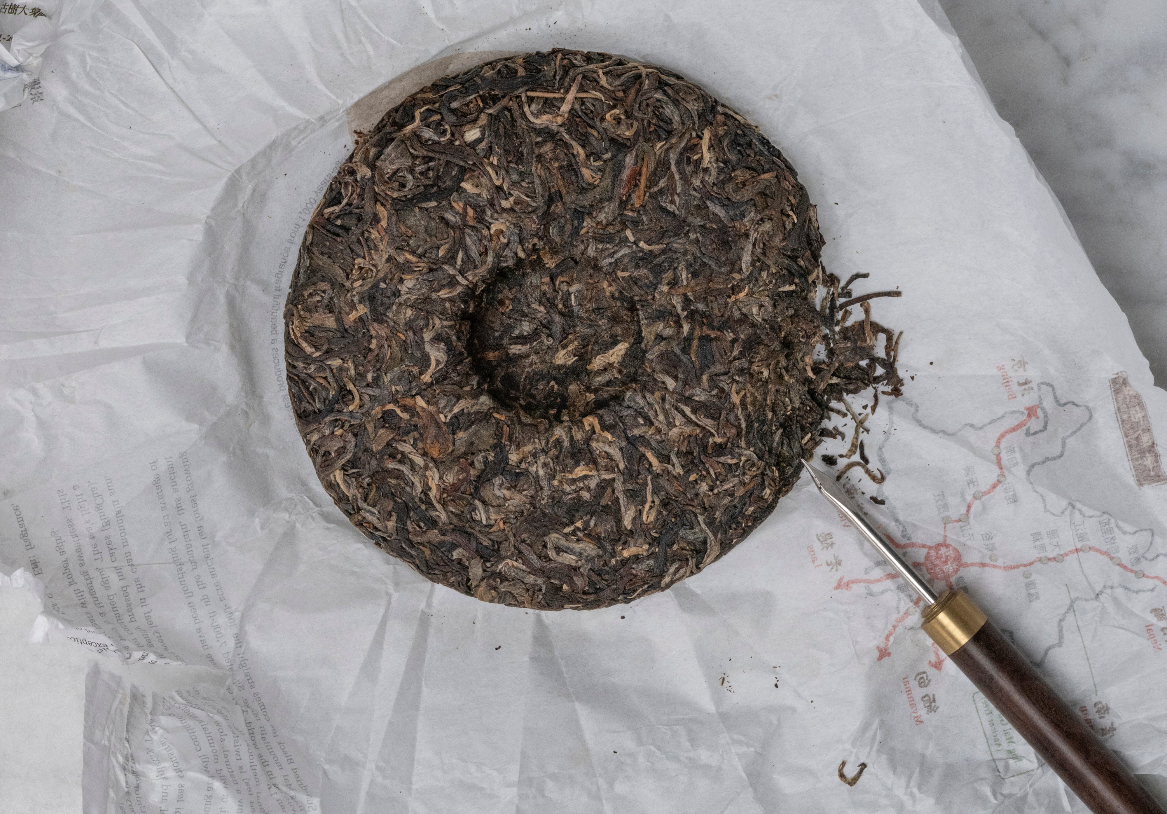 2014 Jingmai Old Forest pu erh tea cake, unwrapped and resting on its paper with a tea needle next to a few leaves picked off the cake.