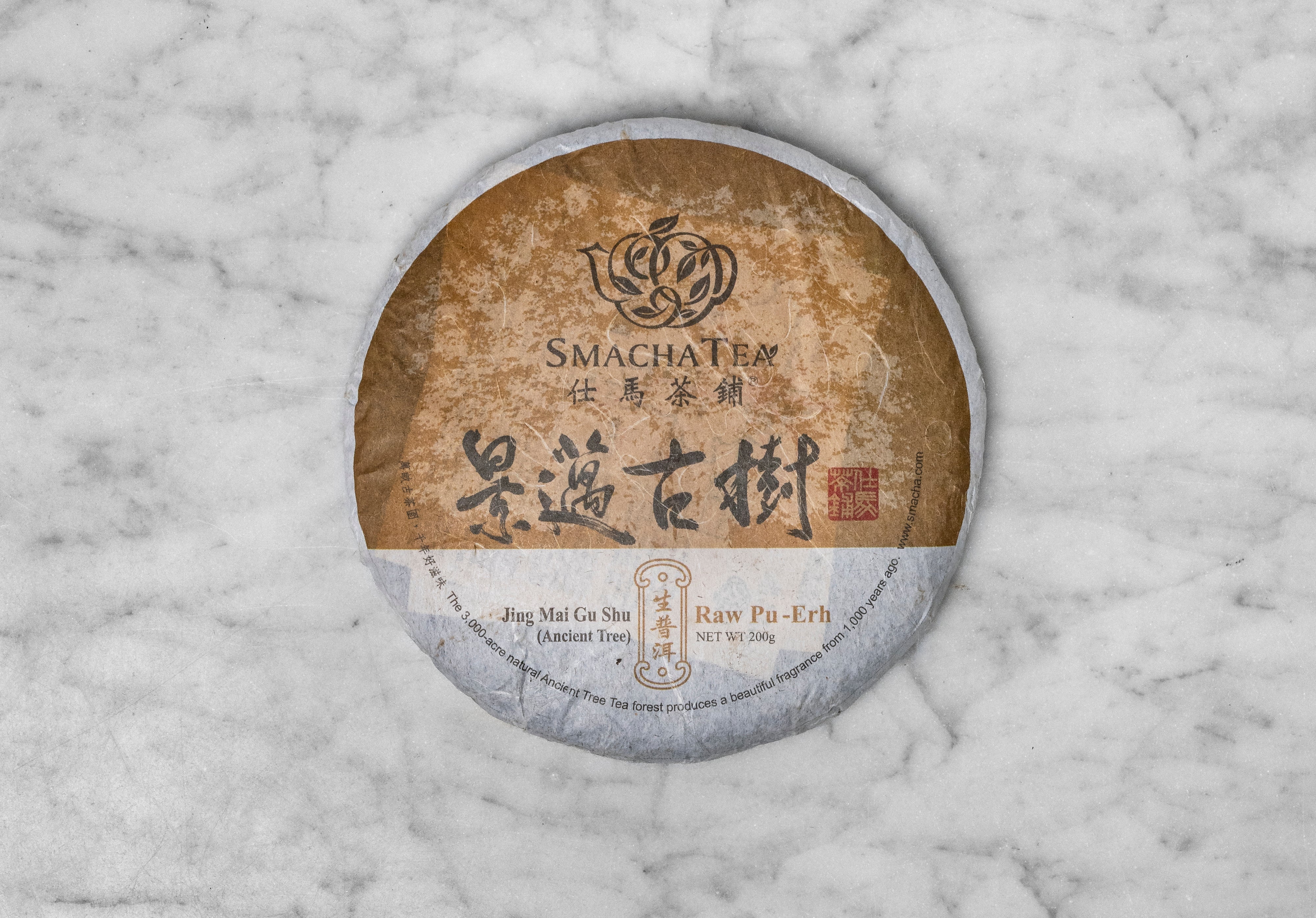 2014 Jingmai Old Forest shen pu erh tea cake wrapped in paper with the words "Jing Mai Gu Shu (Ancient Tree) Raw Pu Erh".