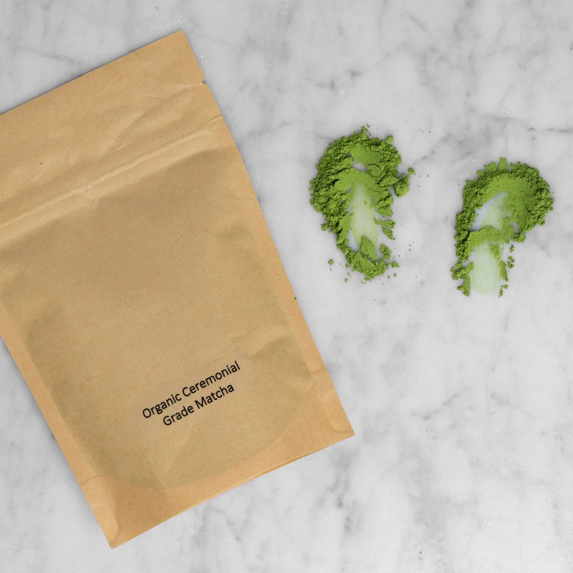 Kraft paper bag labeled 'Organic Ceremonial Grade Matcha' with matcha powder on a marble surface. The matcha is pulled down on the marble to show the fine quality of the stone ground matcha. 