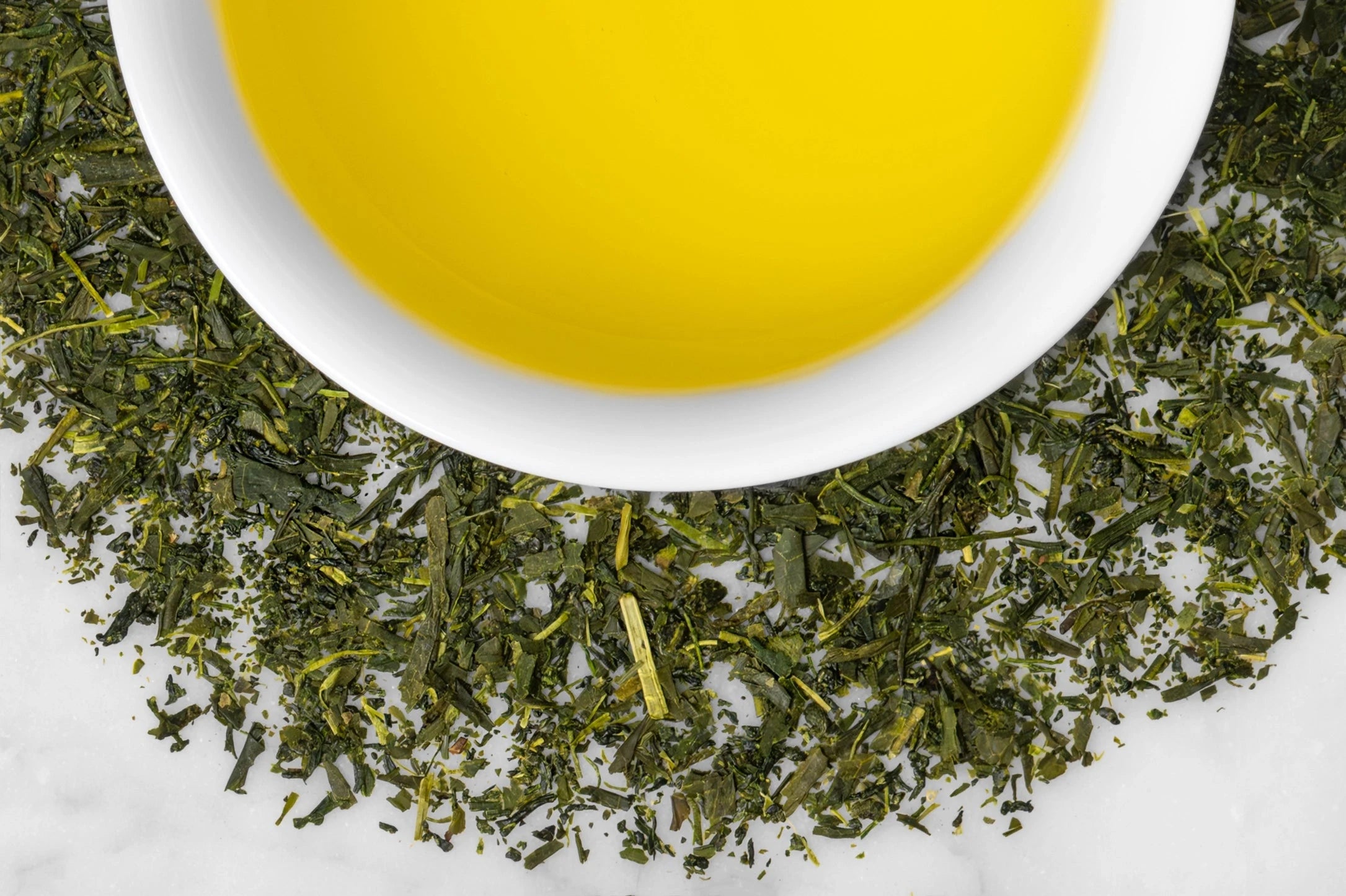 Organic Sencha Fukamushi leaves and brewed tea. Thin, dark green Sencha leaves of varying sizes surround a cup of bright yellow-green brewed tea.