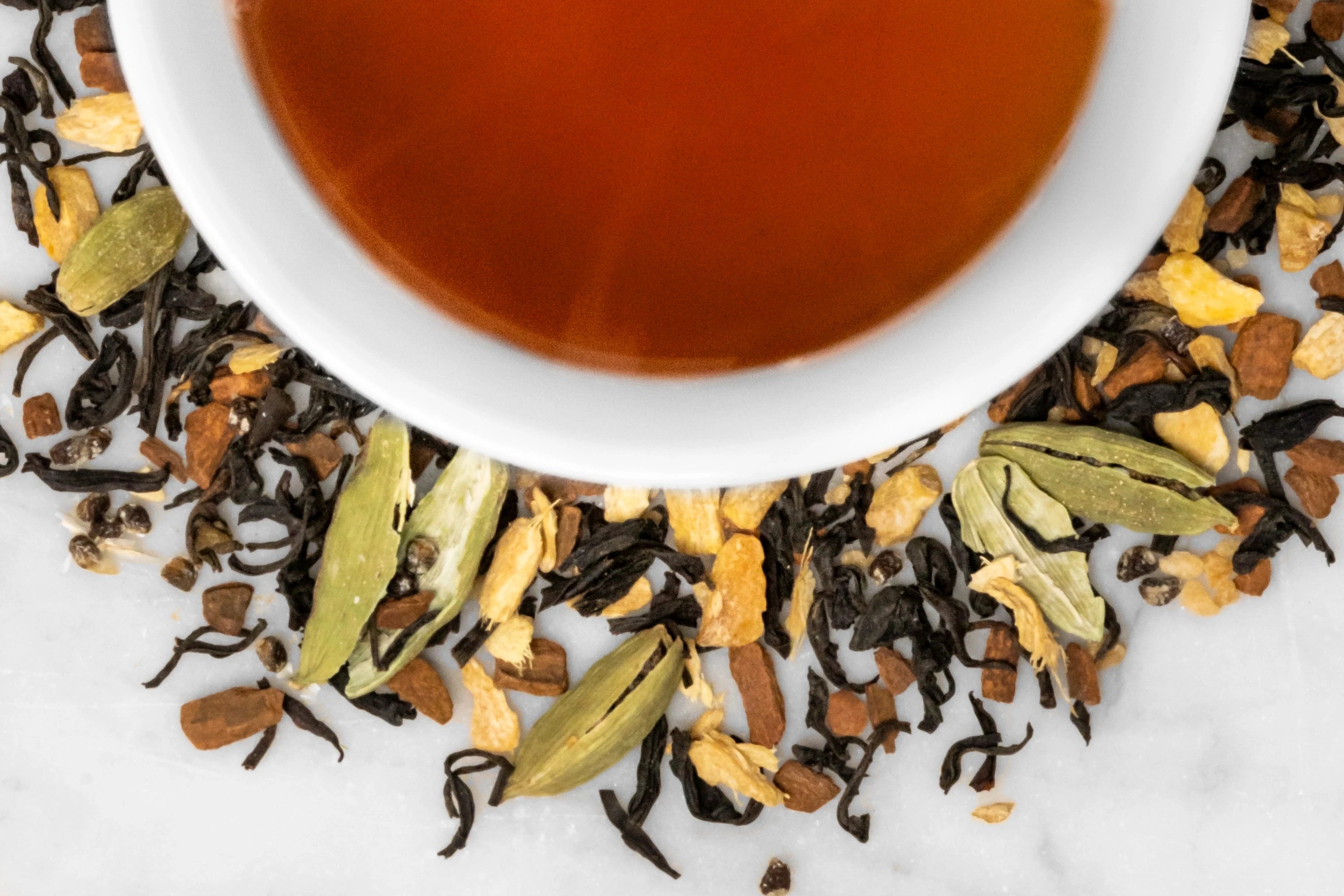 Close up of dry and brewed Pantry Hill Blend. The dry tea is composed of black tea leaves, ginger, crushed cardamom pods, and cinnamon around a white cup with dark reddish-brown brewed tea.