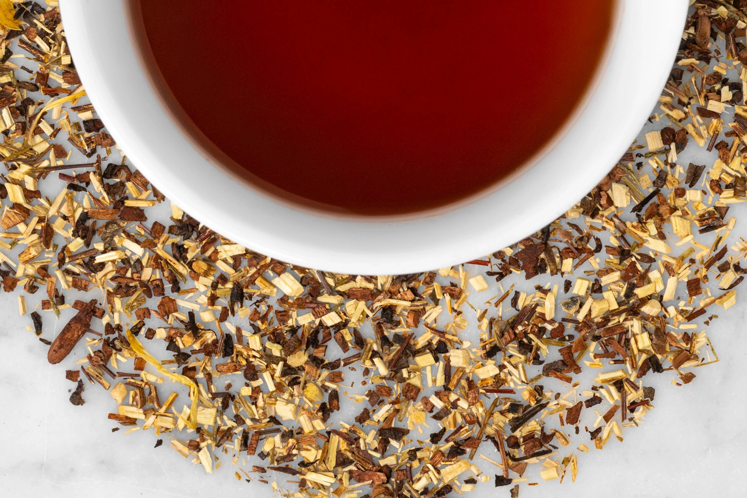 Brewed and dry Nana's Pumpkin Pie herbal tisane. A dark, reddish-brown cup of brewed tea is surrounded by small loose leaf rooibos, ginger, honeybush, clove, and nutmeg. 