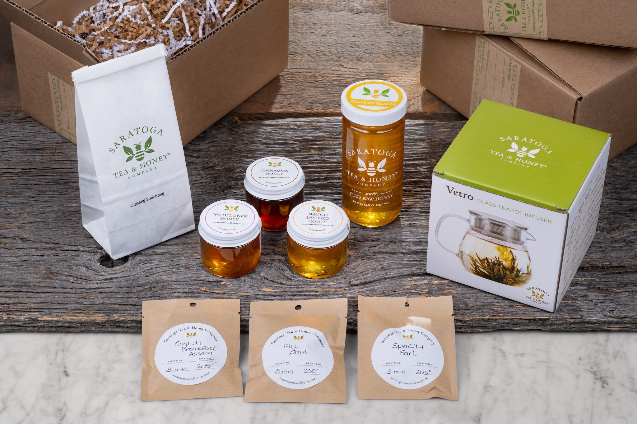 flat lay of queen bee gift set featuring three tea samples, three honey samples, a glass tea pot, tea scoop, 2oz bag of tea, 12 oz jar of european honey, and a local dark chocolate and tea bar with shipping boxes