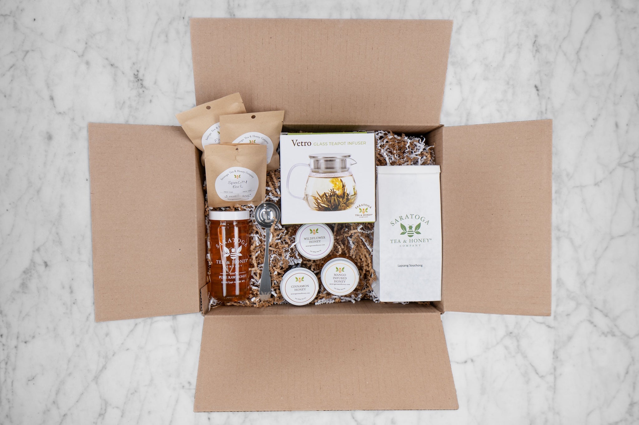 open subscription box with decorative fill, tea samples, mini honeys large honey, large tea, tea scoop, and glass teapot