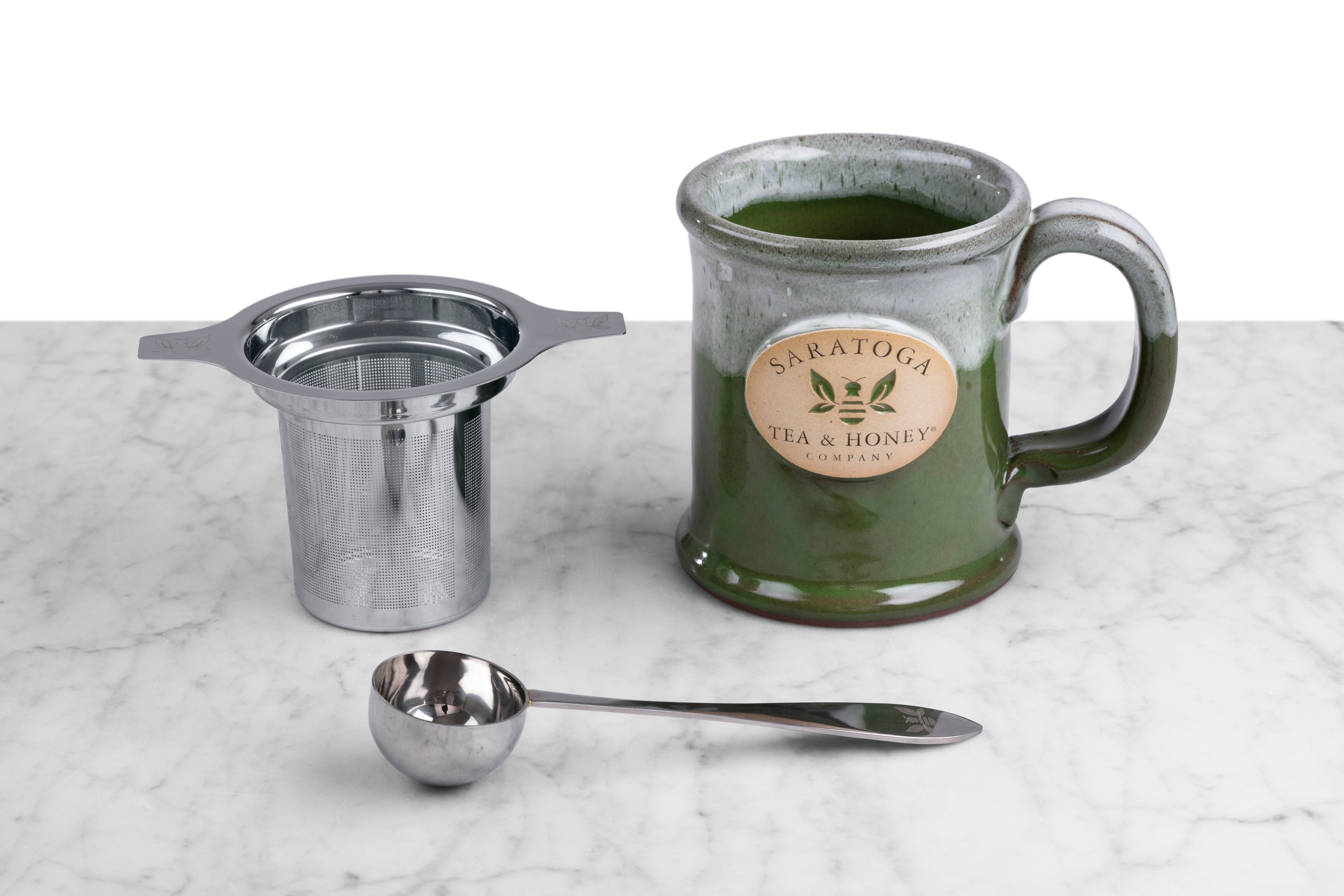 stainless steel tea infuser, tea scoop, and saratoga tea and honey co branded mug on a marble and white background