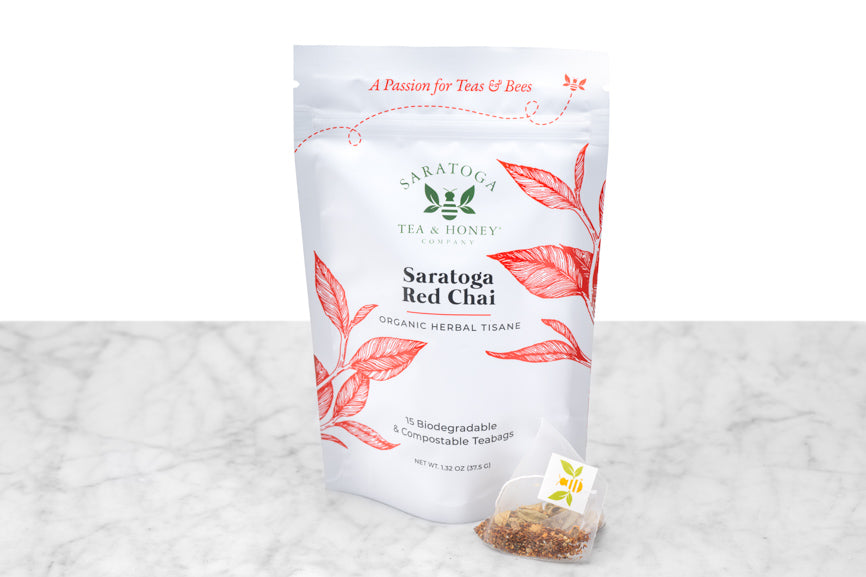 Saratoga Red Chai tea sachet packaging and sample tea sachet. Small, pyramid-shaped tea sachet with visible tea and a bee-logo tag sits next to a pouch reading "Saratoga Red Chai Organic Herbal Tisane. 15 Biodegradable & Compostable Teabags."