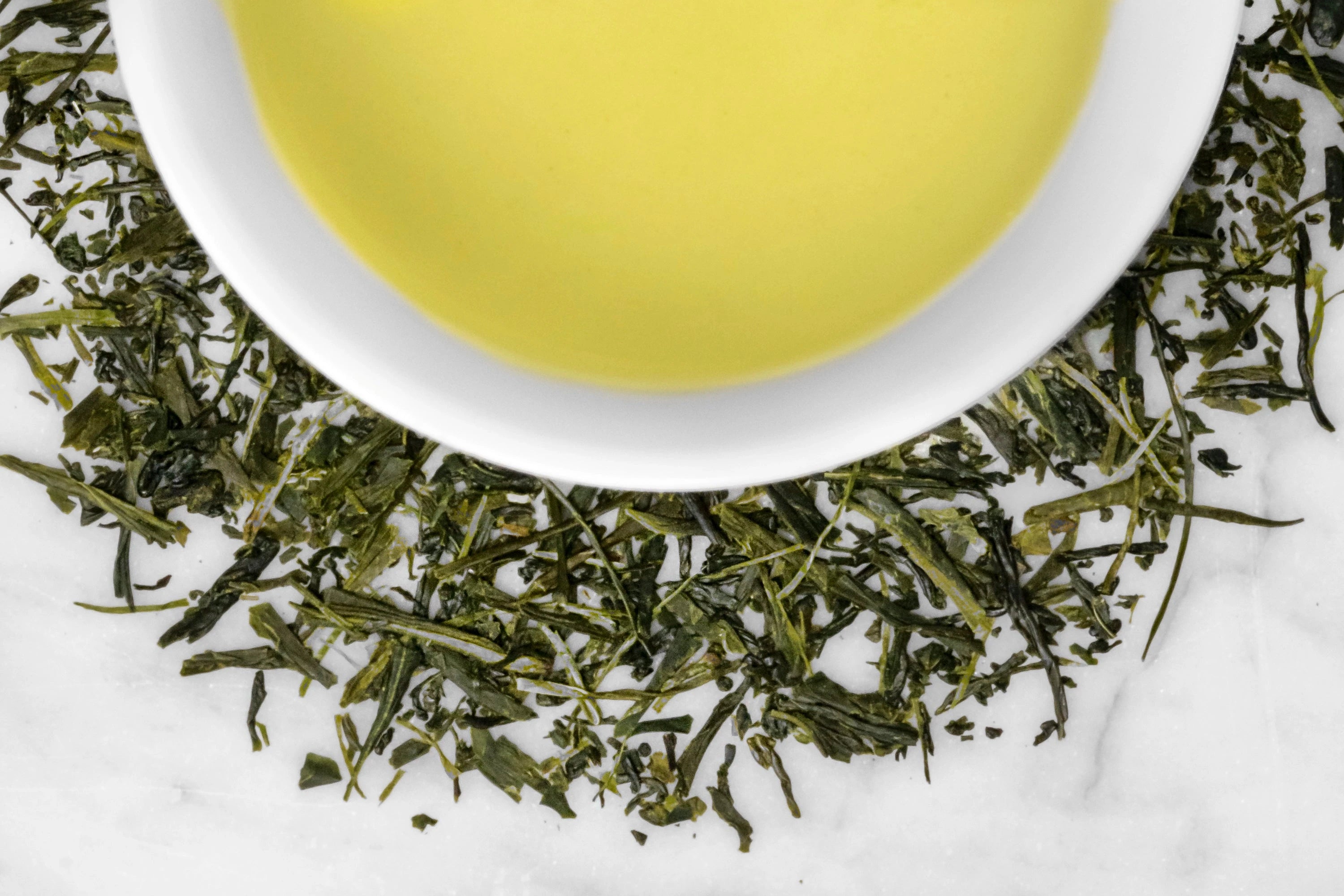 Organic Sencha Ashikubo leaves and brewed tea. Thin, dark green Sencha leaves of varying sizes surround a cup of bright yellow-green brewed tea.