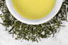 Organic Sencha Ashikubo leaves and brewed tea. Thin, dark green Sencha leaves of varying sizes surround a cup of bright yellow-green brewed tea.