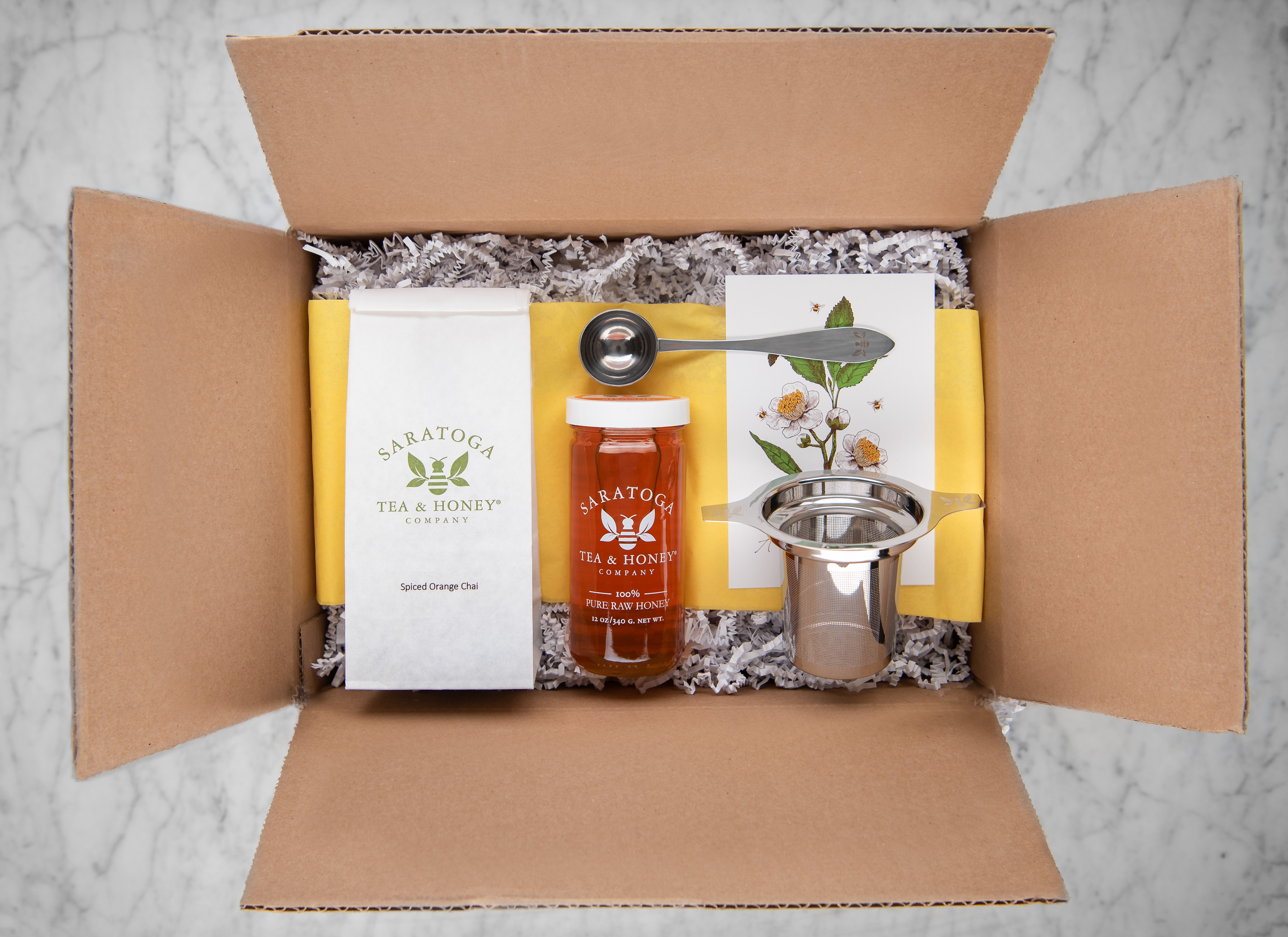 winter tea and honey box: cardboard box with decorative fill, spiced orange chai, jar of orange blossom honey, tea scoop and tea infuser