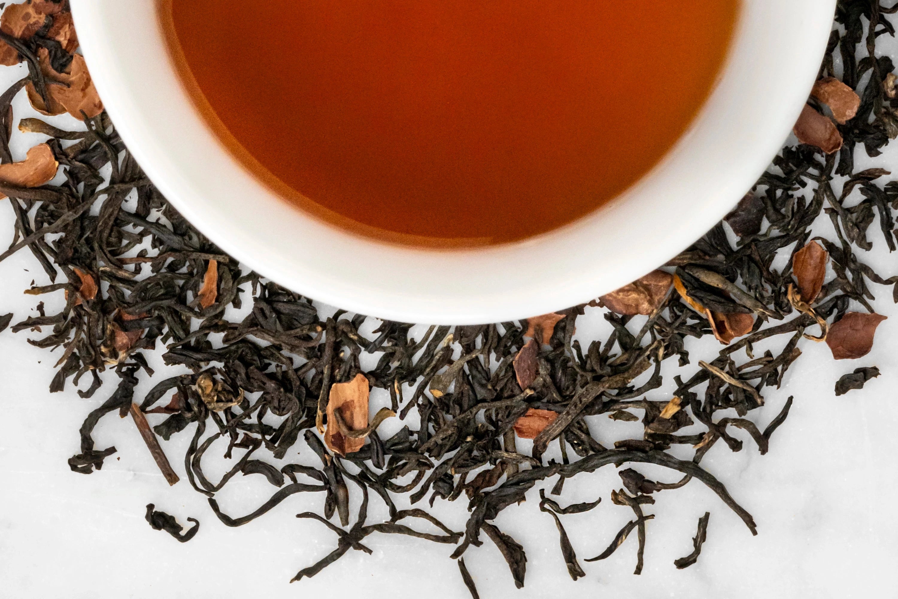Close up of dry and brewed Tea Quiero. The dry tea is composed of black tea leaves and cacao pieces around a white cup with dark reddish-brown brewed tea.