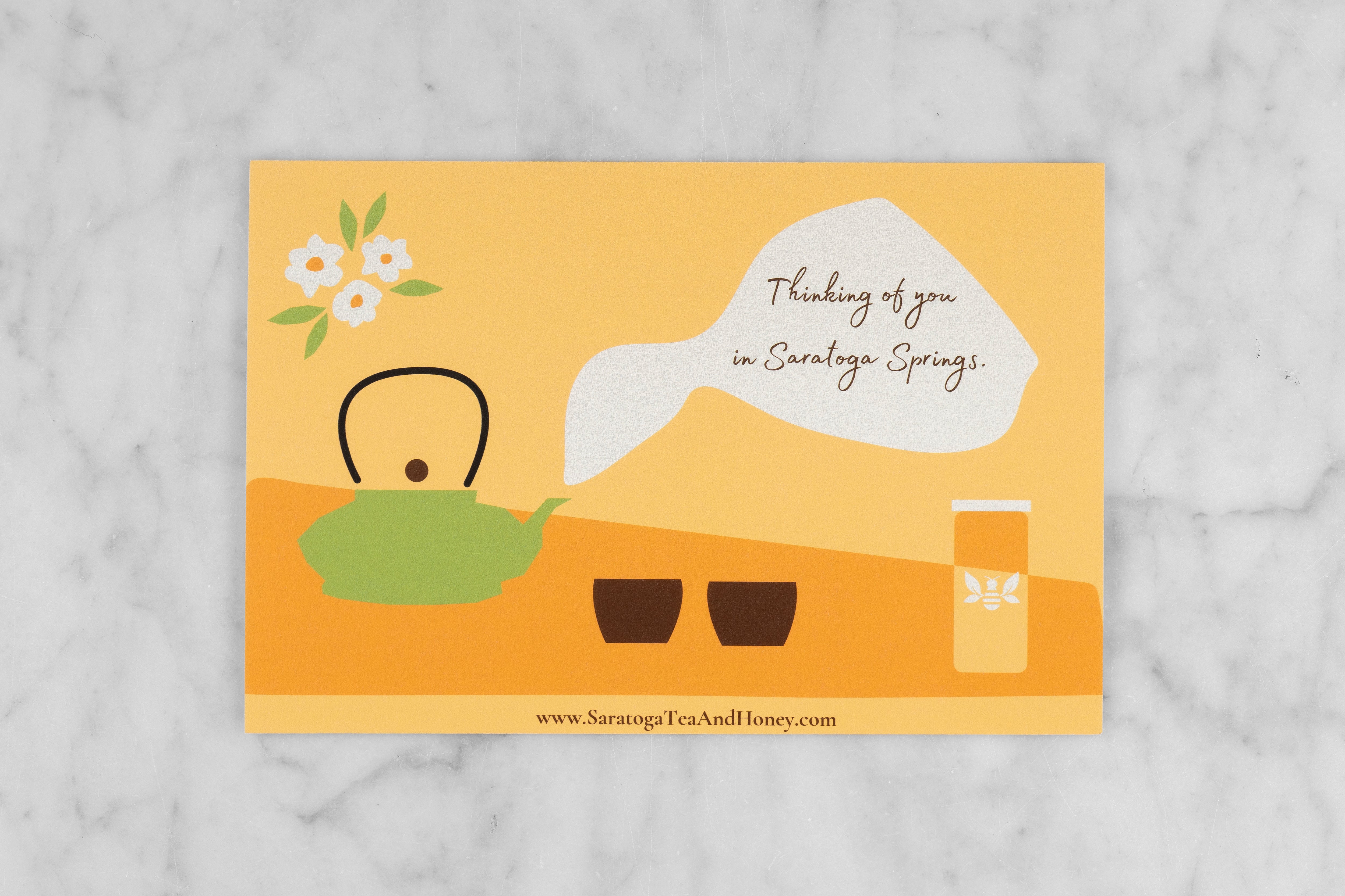 yellow postcard with green teapot and cups and the message: "Thinking of you in Saratoga Springs." on a marble background