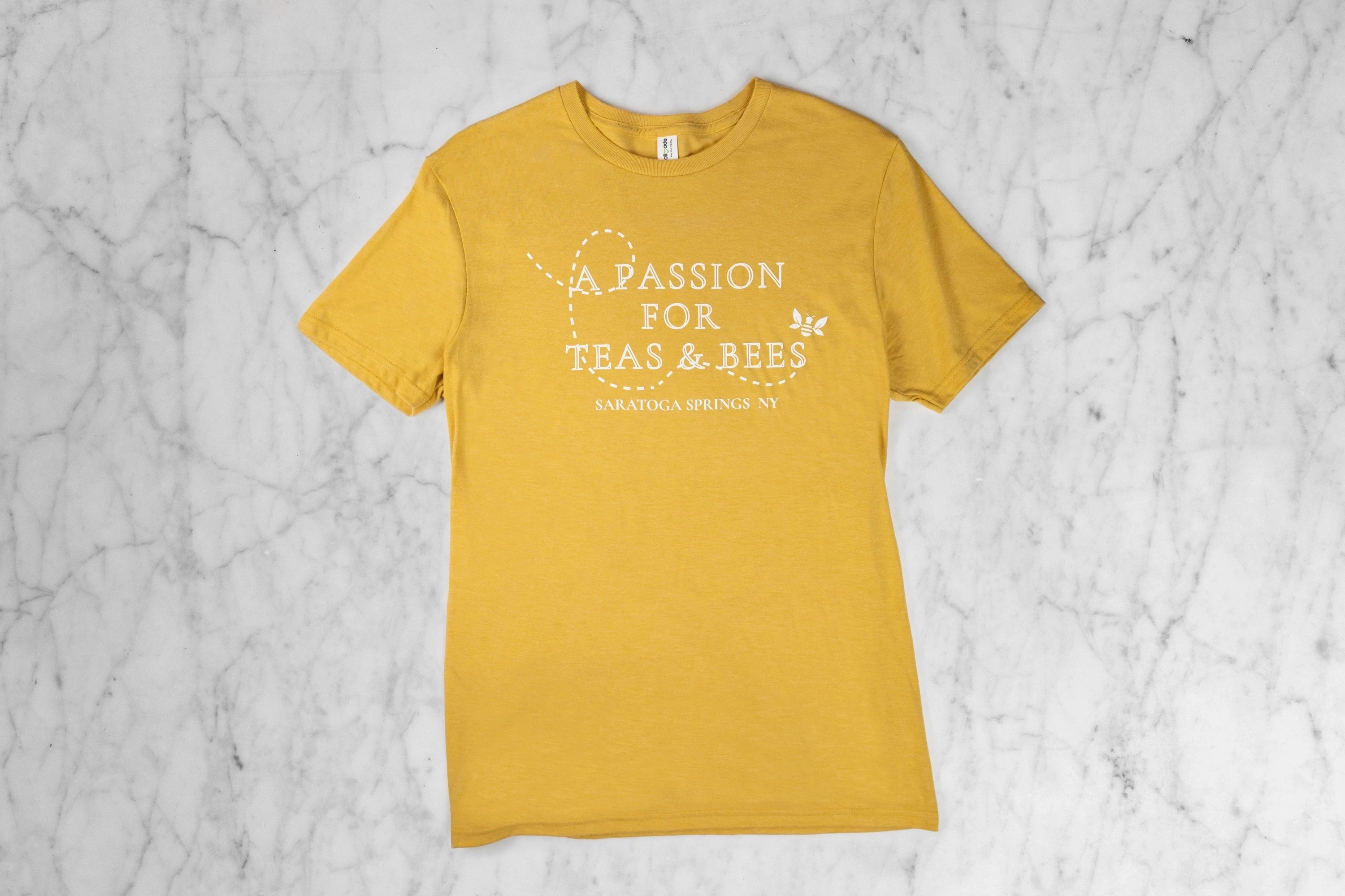 Sustainable T-Shirt Front. Yellow unisex T-Shirt with the words "A Passion for Teas & Bees Saratoga Springs NY" and a Saratoga Tea & Honey Co. Tea Bee flying with a dotted swirly line showing its path.